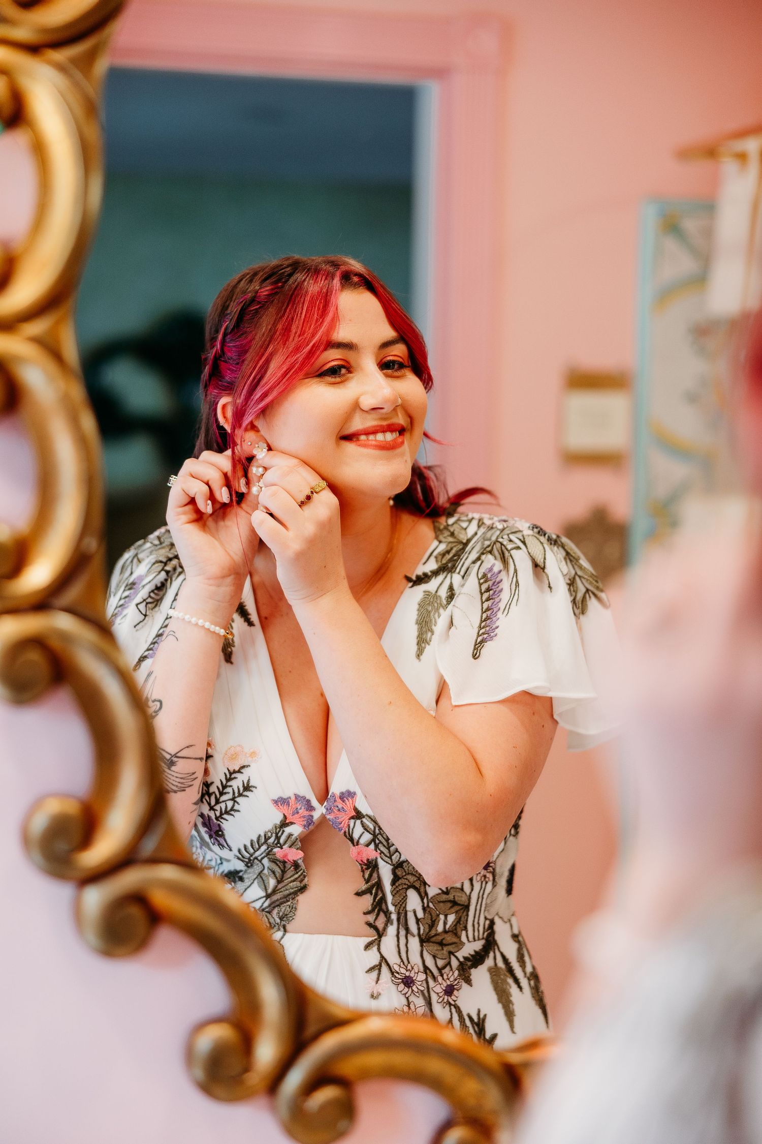 A reflection in an ornate gold mirror shows someone with vibrant red hair and a floral dress smiling warmly.