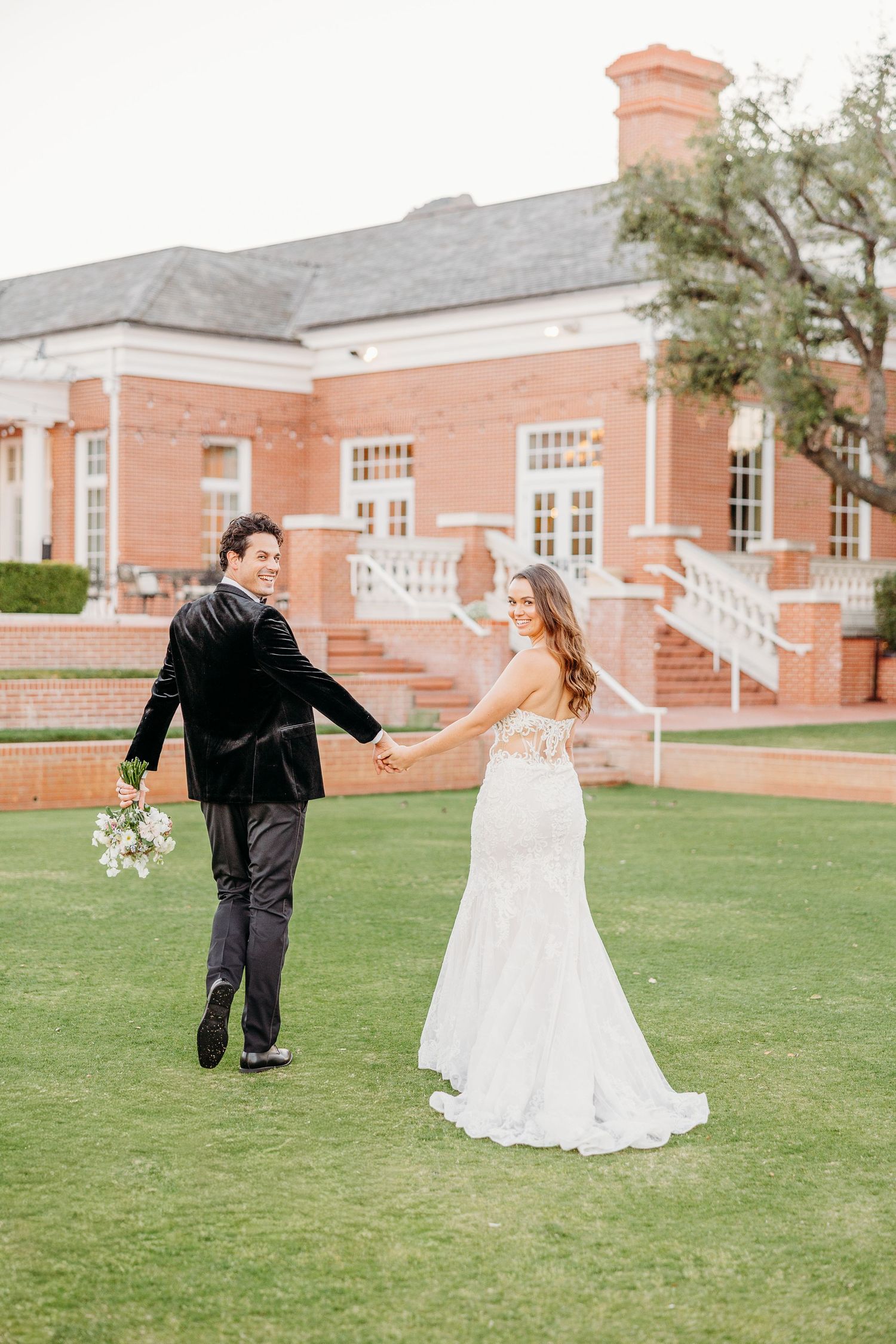 A couple runs playfully on the lawn in front of a pink brick building during their wedding celebration.