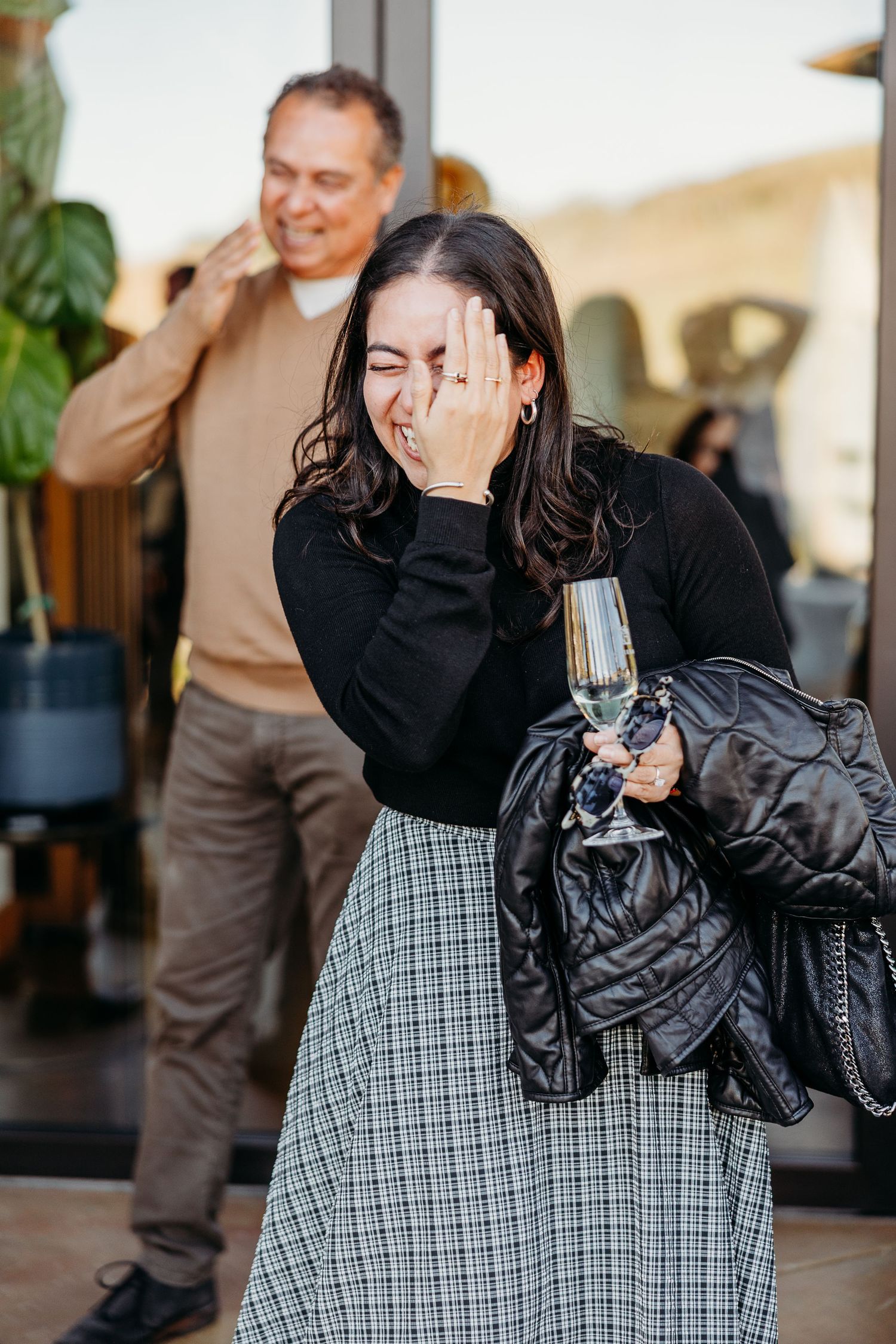 A person in a black sweater and plaid skirt covers their face in surprise while holding a wine glass.
