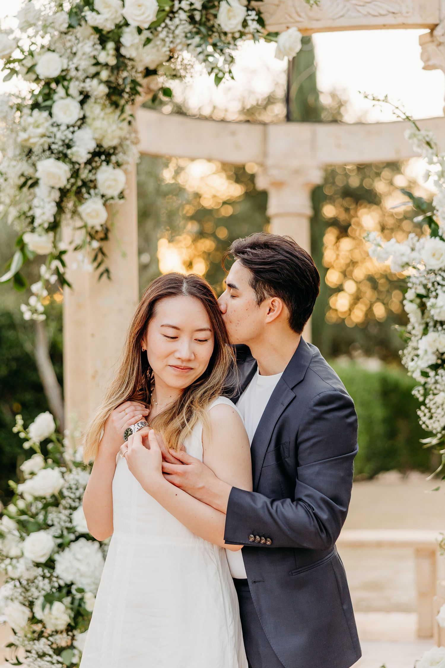 Intimate moment between couple under white floral gazebo during sunset photoshoot.