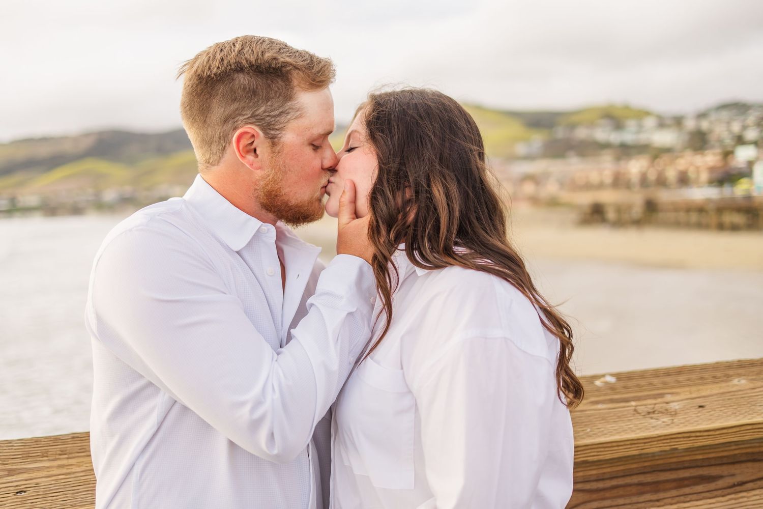 A tender moment between a couple in white shirts embracing on a pier with a coastal backdrop.