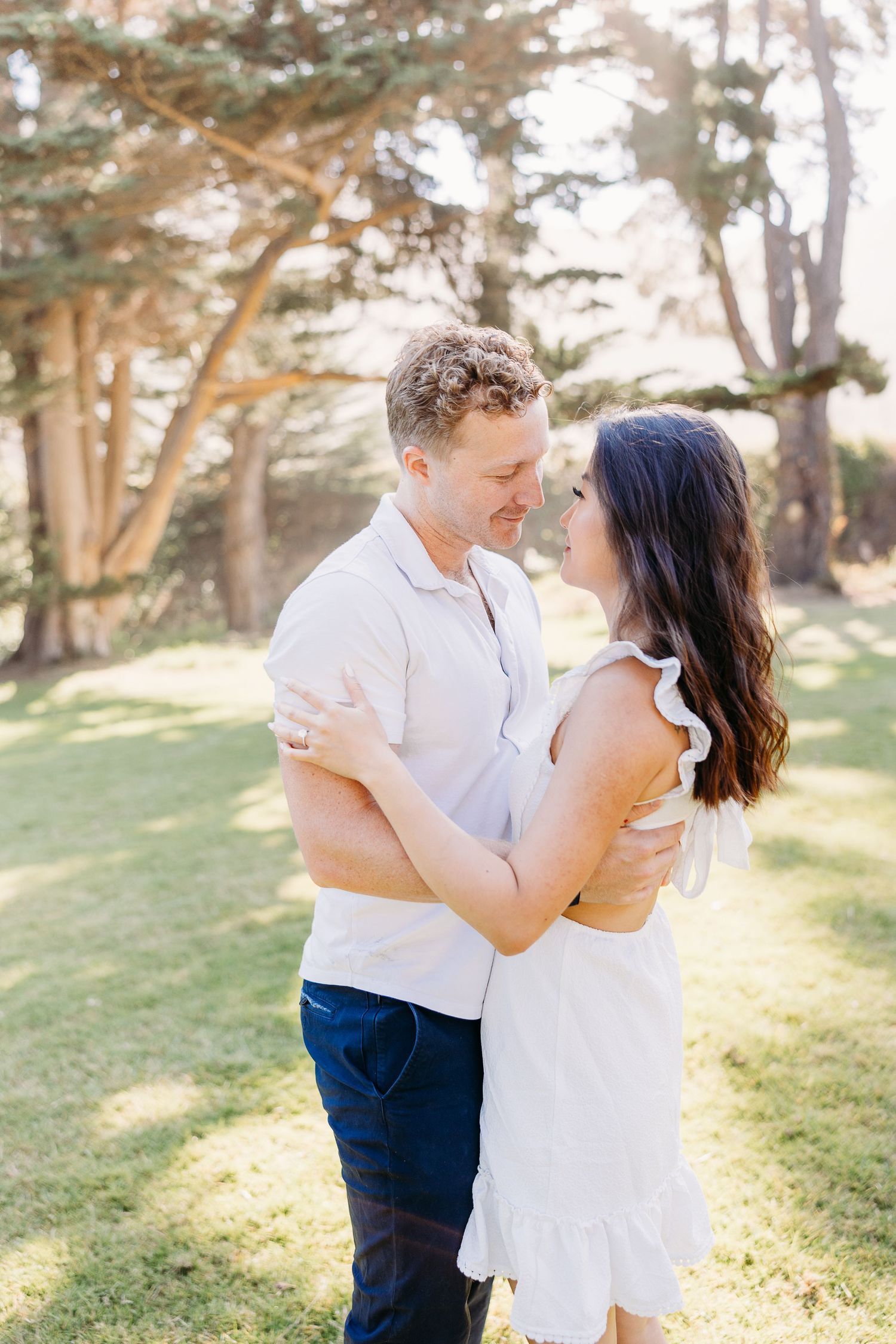 A romantic couple embraces in a sunlit park with tall trees in the background.