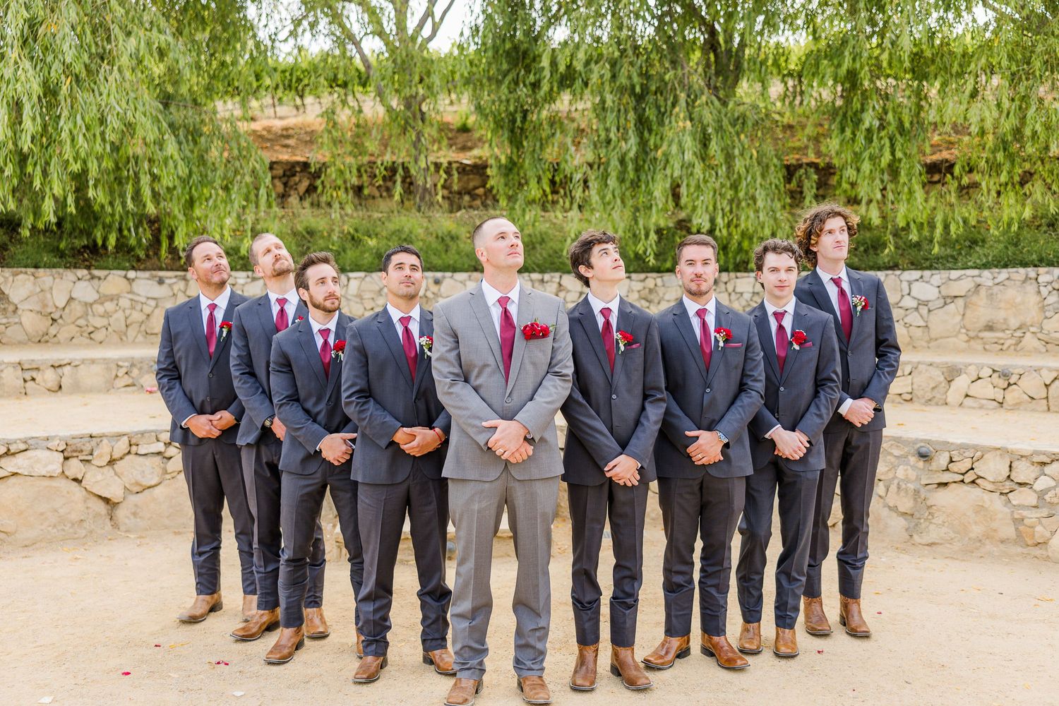 Groomsmen in dark suits with burgundy ties pose on stone steps under willow trees.
