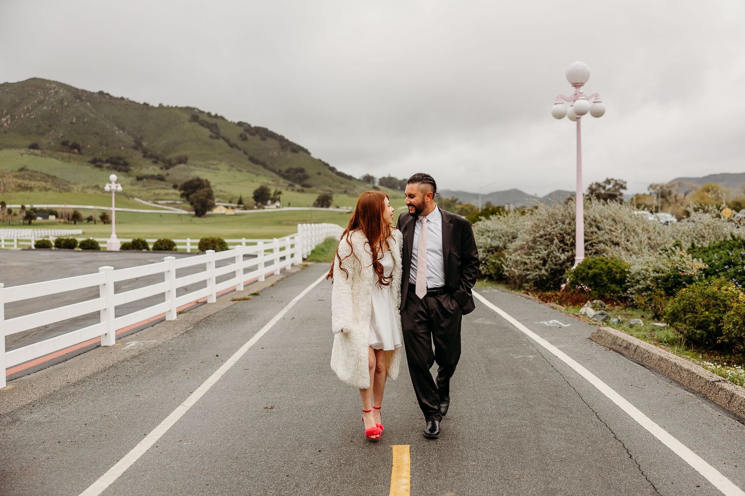 A couple walk hand in hand down a rural road lined with white fences and rolling hills in the background.