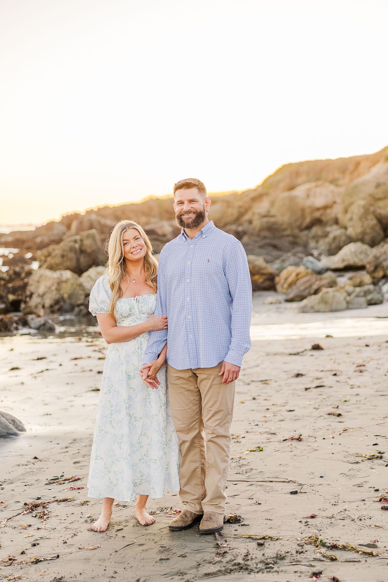 A couple stands together on a sandy beach with coastal cliffs behind them at sunset.