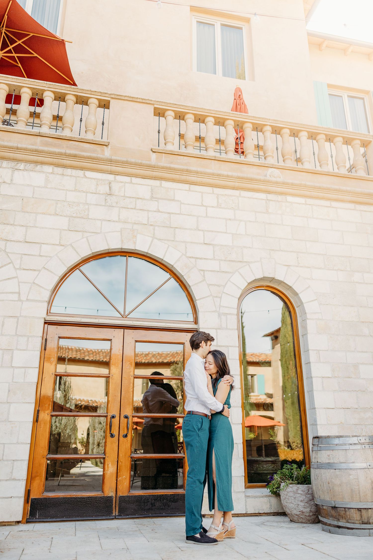 Elegant couple in formal wear share a romantic moment outside a stone building with arched windows.