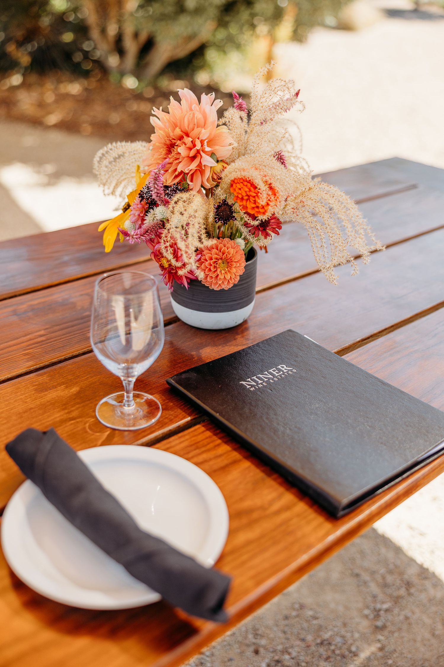 Table setting with wine glasses, black napkins, and coral floral centerpieces on a wooden outdoor table.