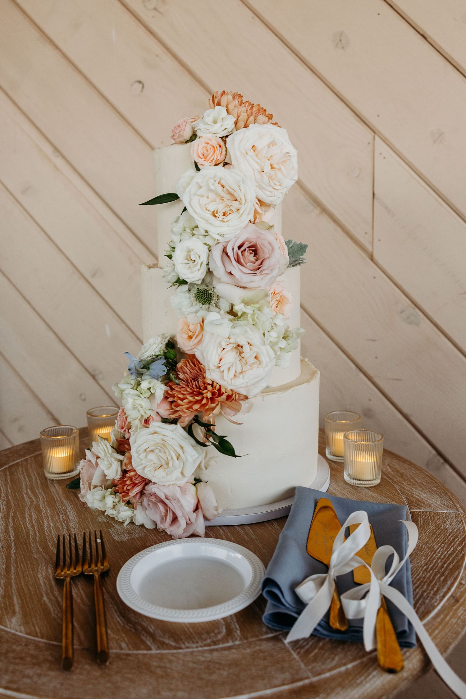 White wedding cake with blush and coral floral decorations on rustic wooden table with place settings and candlelight.