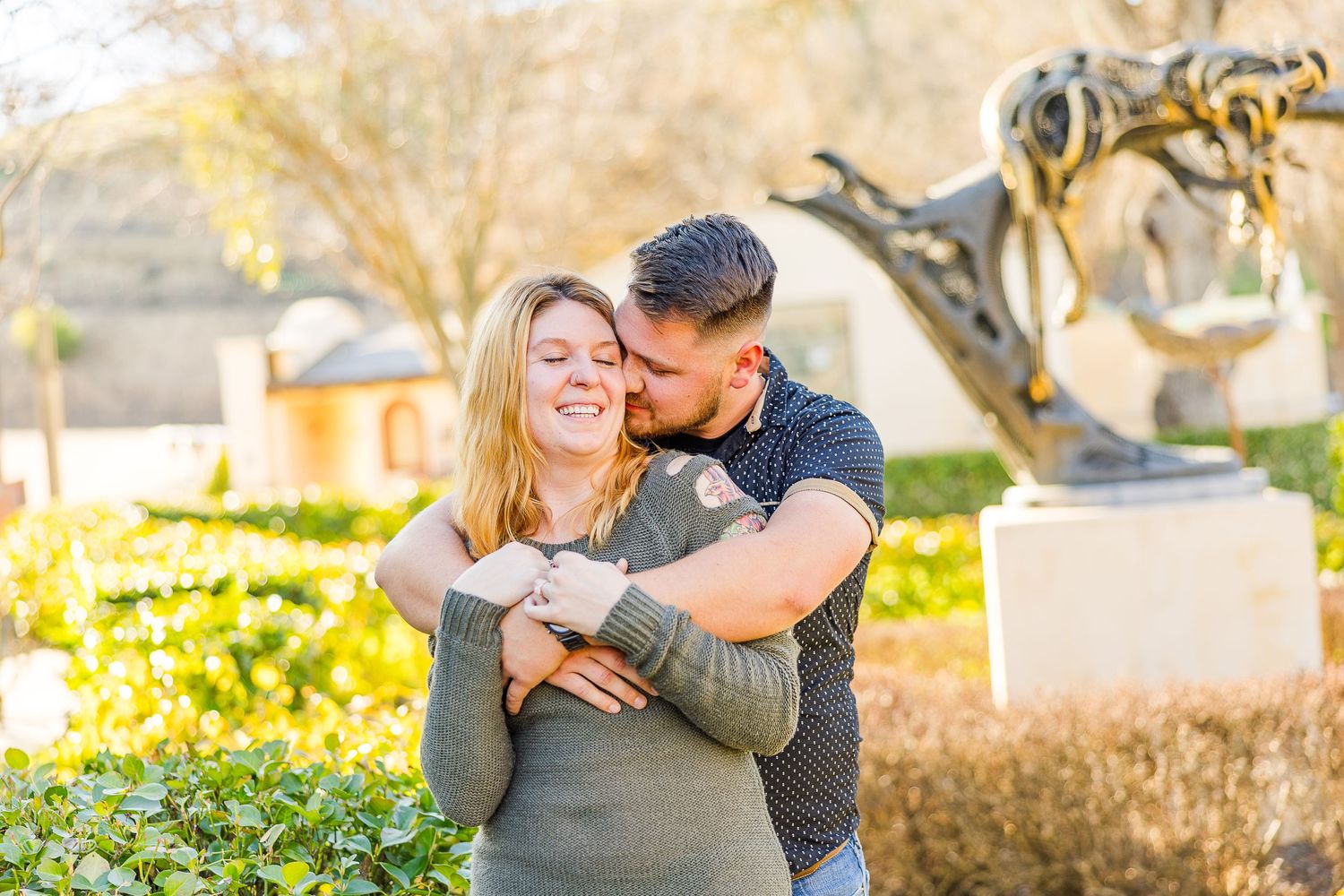 Romantic outdoor photo of couple embracing and laughing in sunlit garden setting.