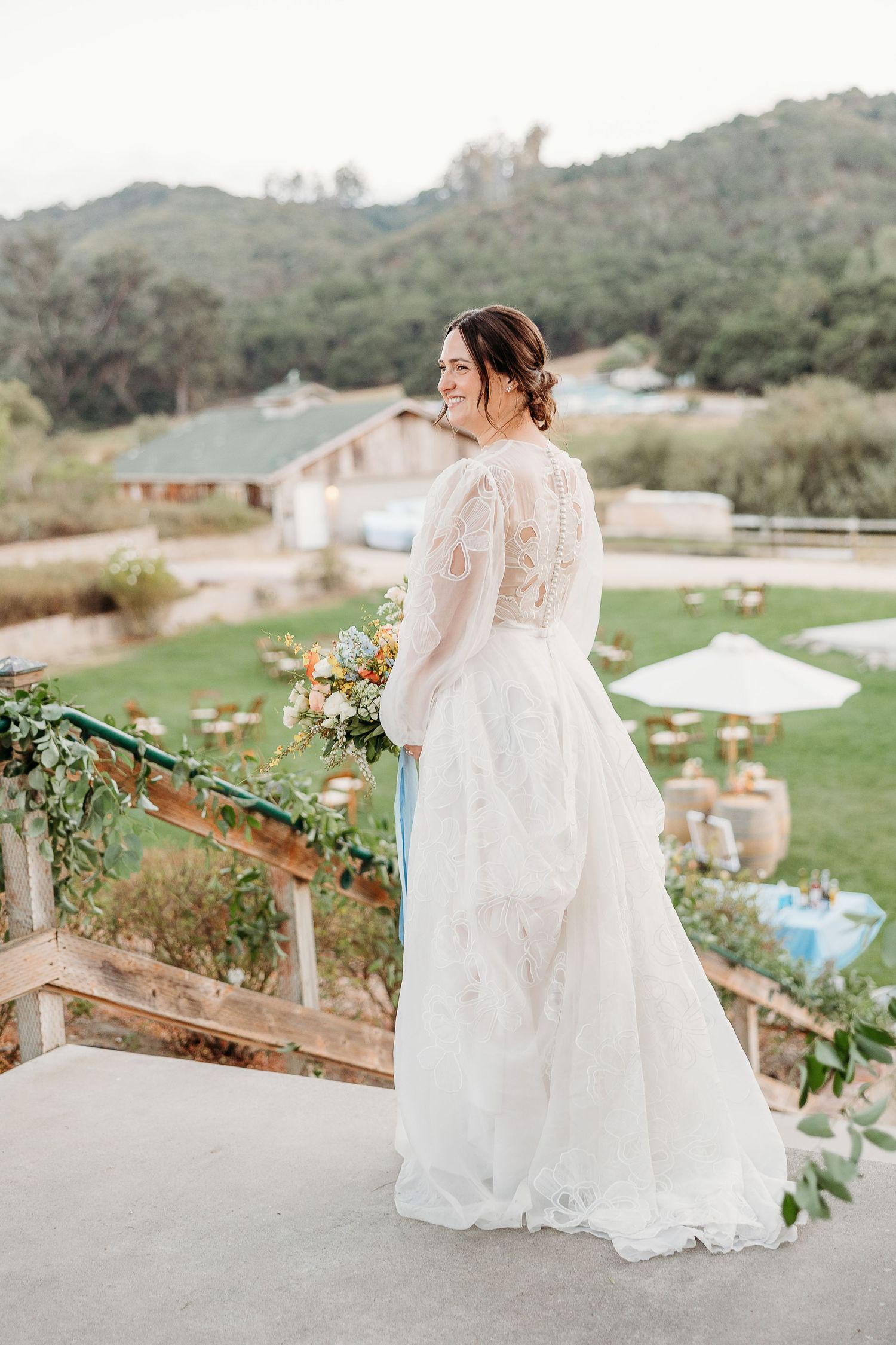Elegant bride in lace wedding dress poses on balcony overlooking mountain venue.