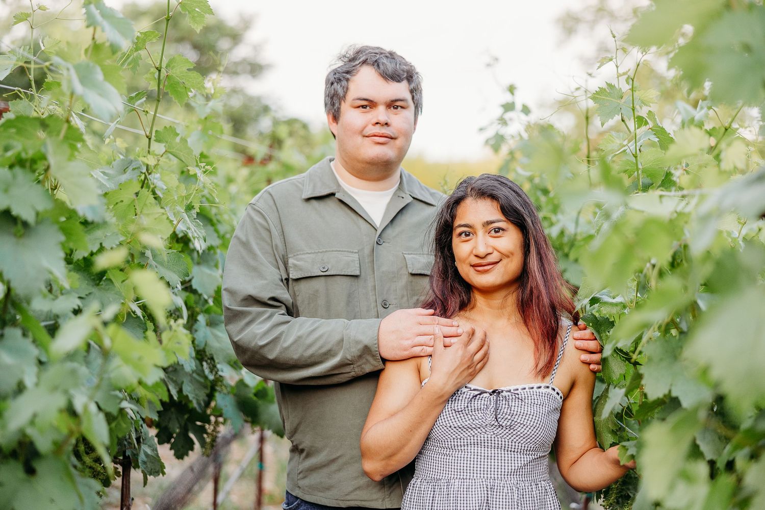 A couple poses together between rows of grapevines in a sunny vineyard setting.
