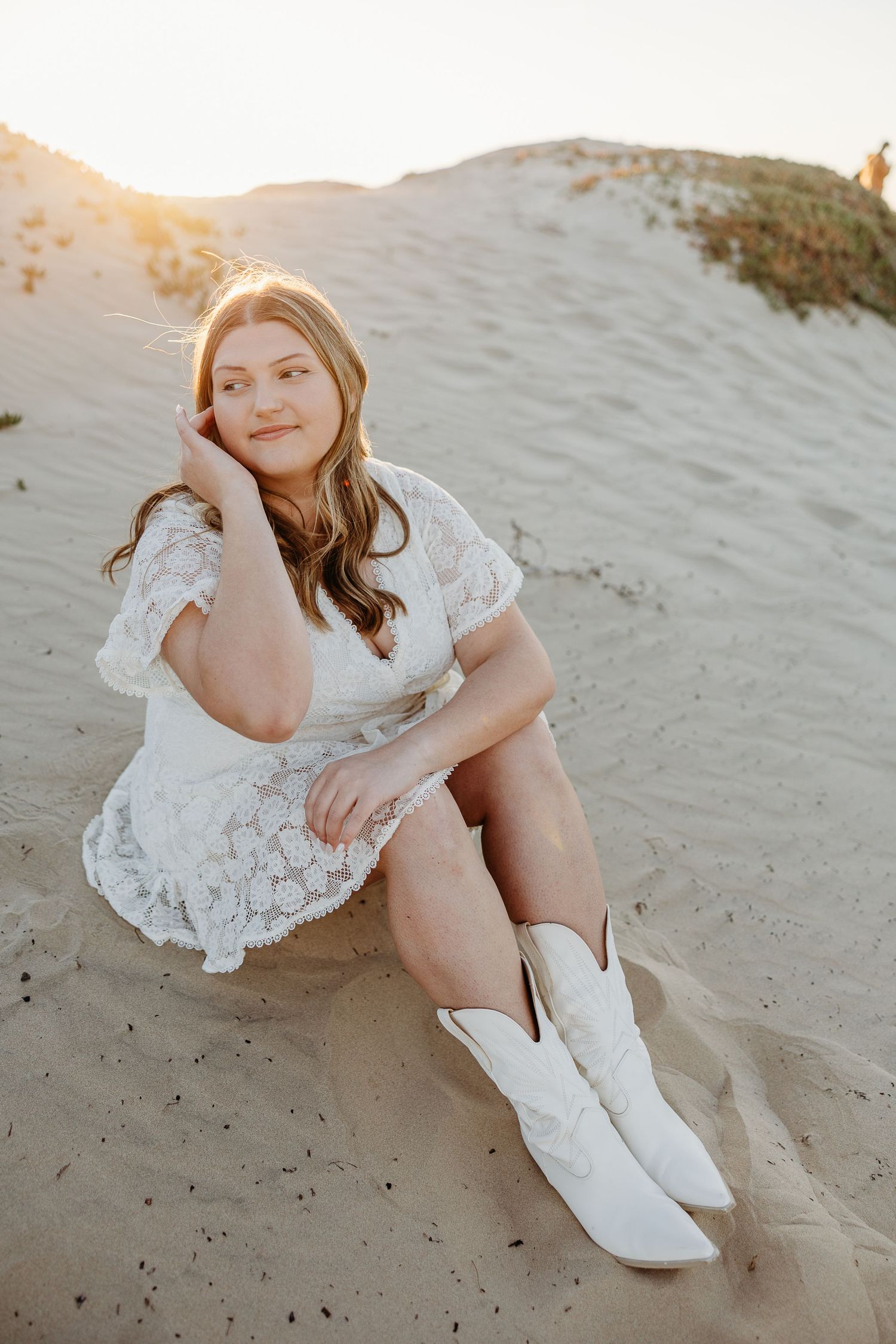 Graduate wearing cowboy boots and lace dress poses elegantly on beach dunes at sunset.