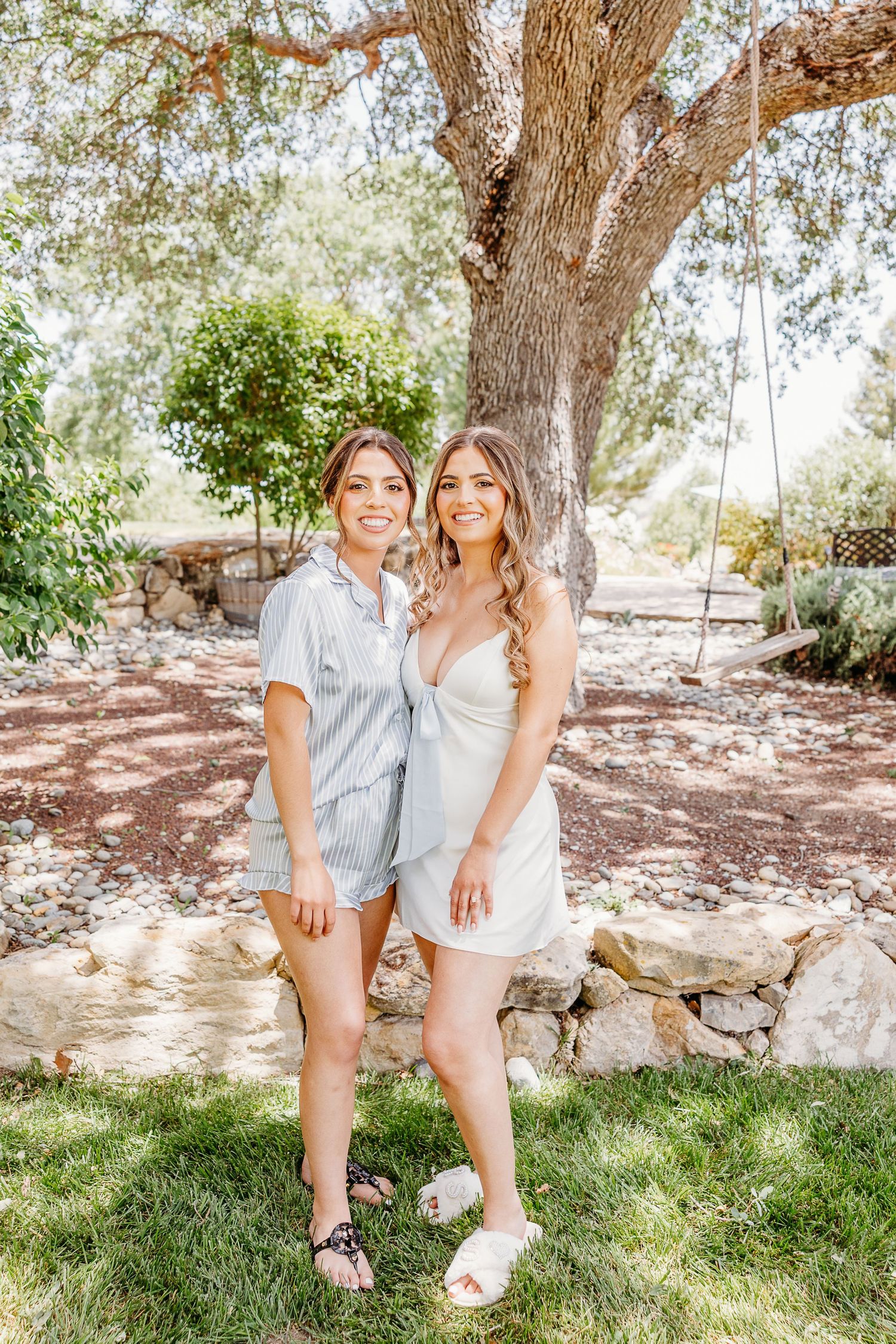 Two people in matching white outfits share joyful moments sitting together under a shady tree on a sunny day.