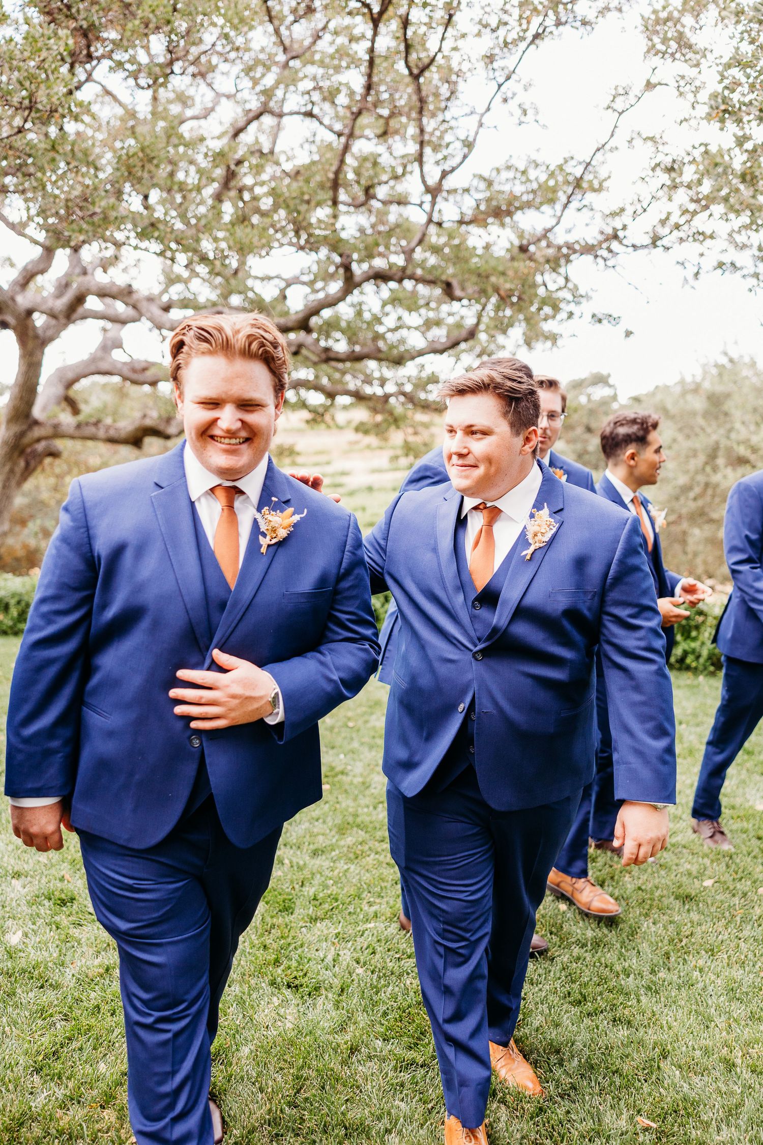 Two groomsmen in bright blue suits and orange ties sharing a laugh outdoors.