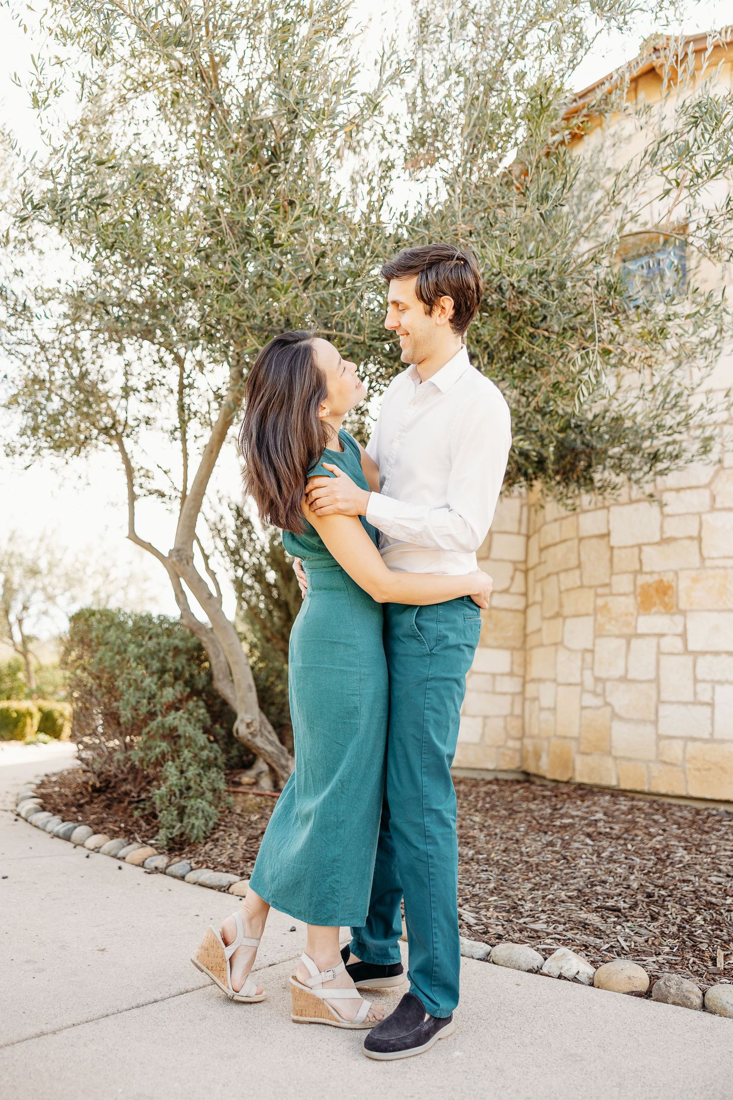 A couple dancing together outdoors near an olive tree and stone building.