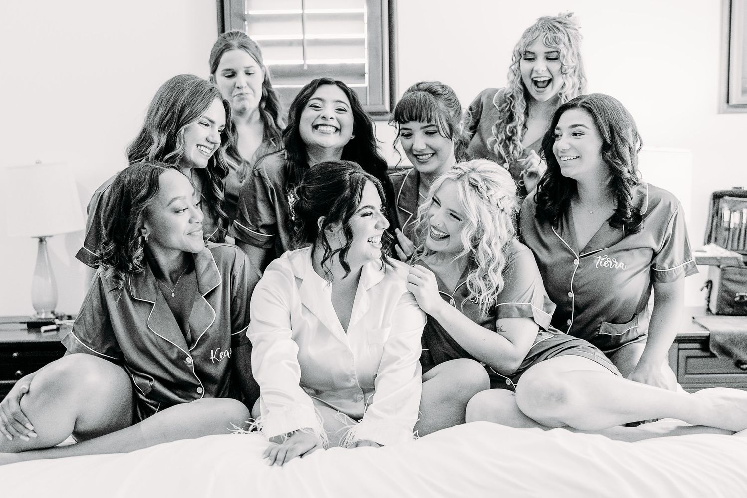Black and white candid photo of a bridal party sharing laughs together on a bed.