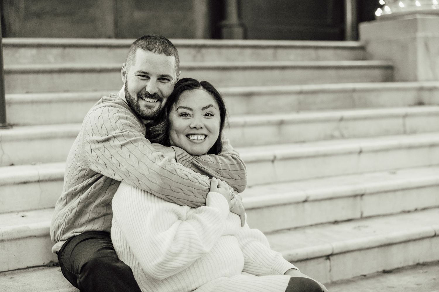 A couple shares a joyful embrace while sitting on outdoor stone steps in black and white.