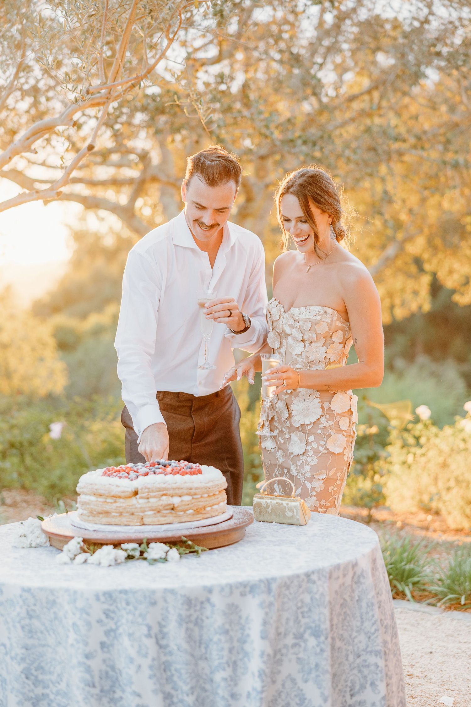 Couple cutting wedding cake together at outdoor reception during sunset.