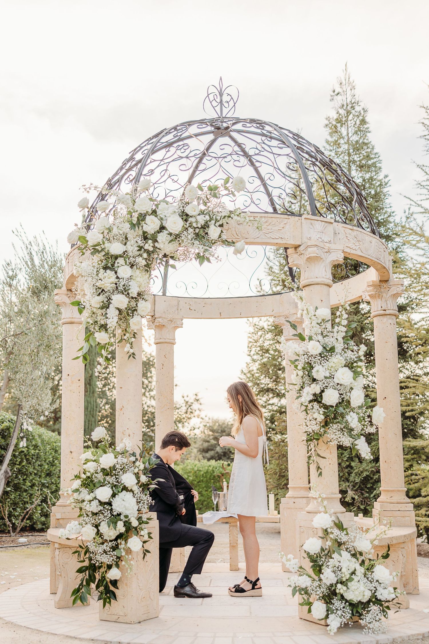Marriage proposal scene under ornate white floral gazebo with classical architecture.