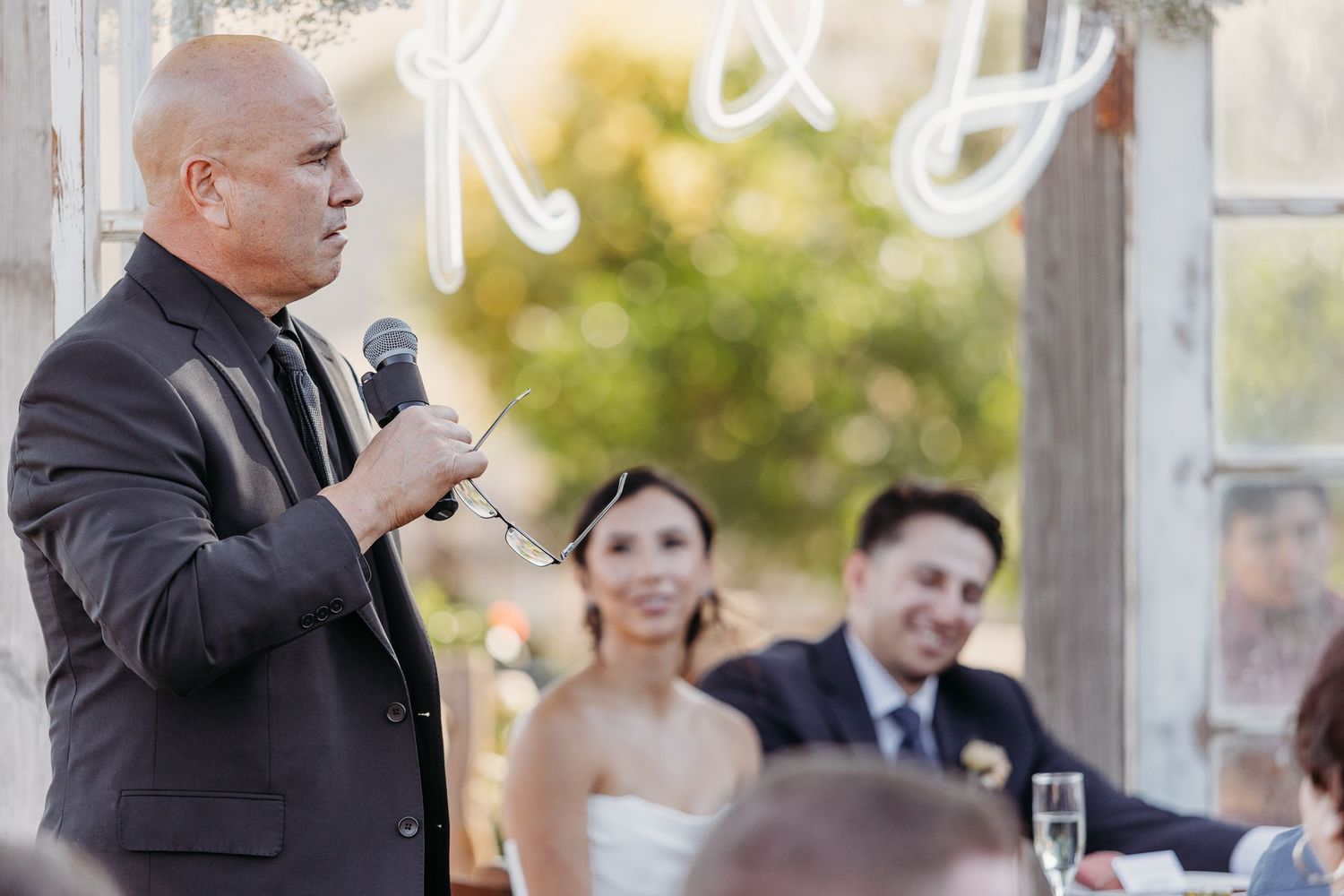 Wedding reception speeches and toasts being given under a clear tent with decorative lighting.