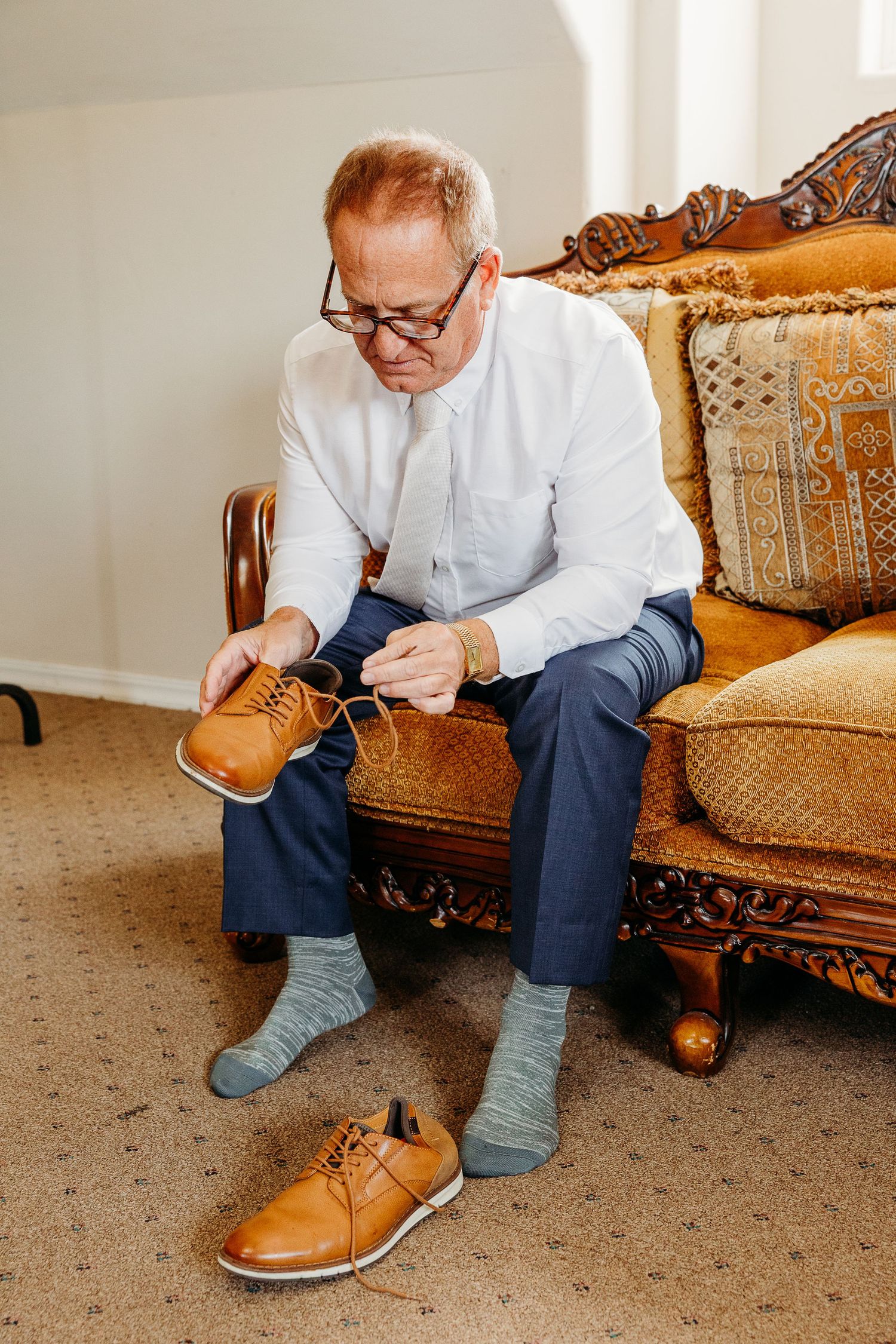 Person putting on brown leather dress shoes while sitting on an ornate vintage couch.
