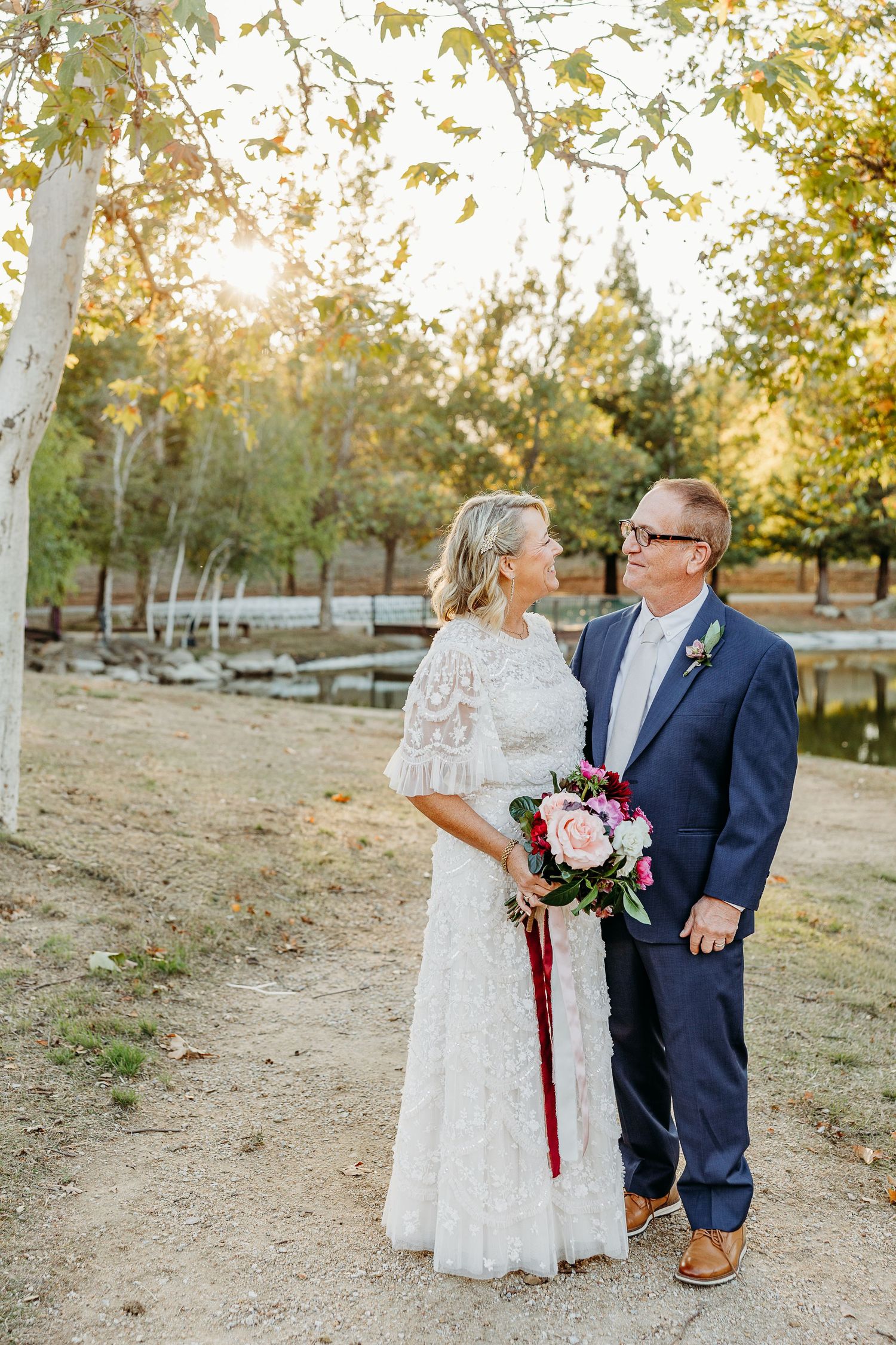 Wedding couple poses together on dirt path with autumn trees in background.