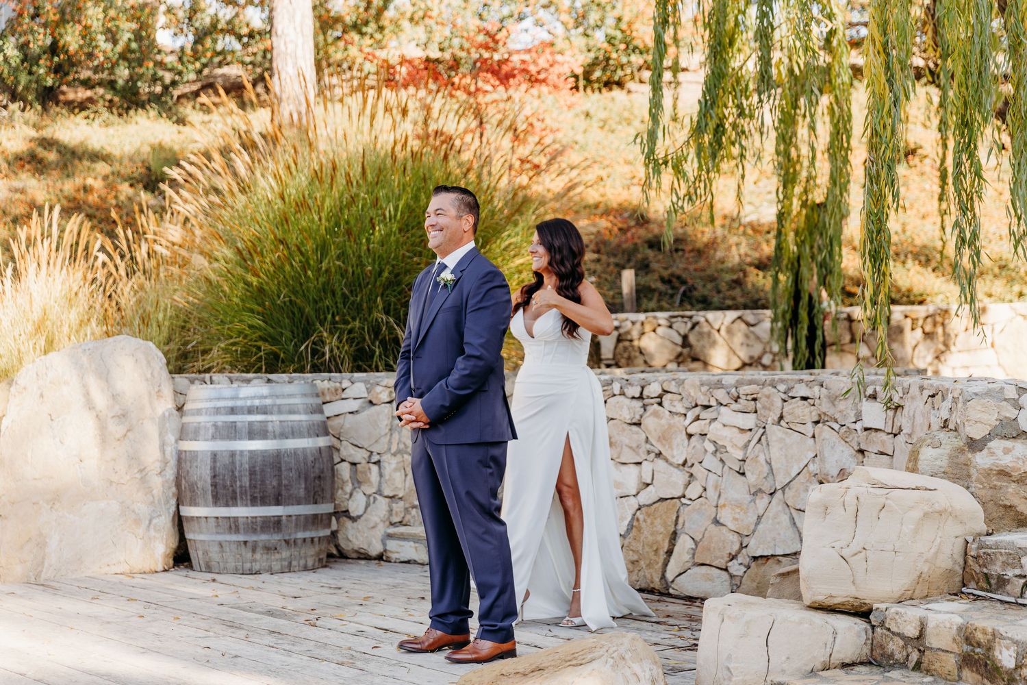 A couple stands at a winery with stone walls, wine barrels and willow trees in the background.