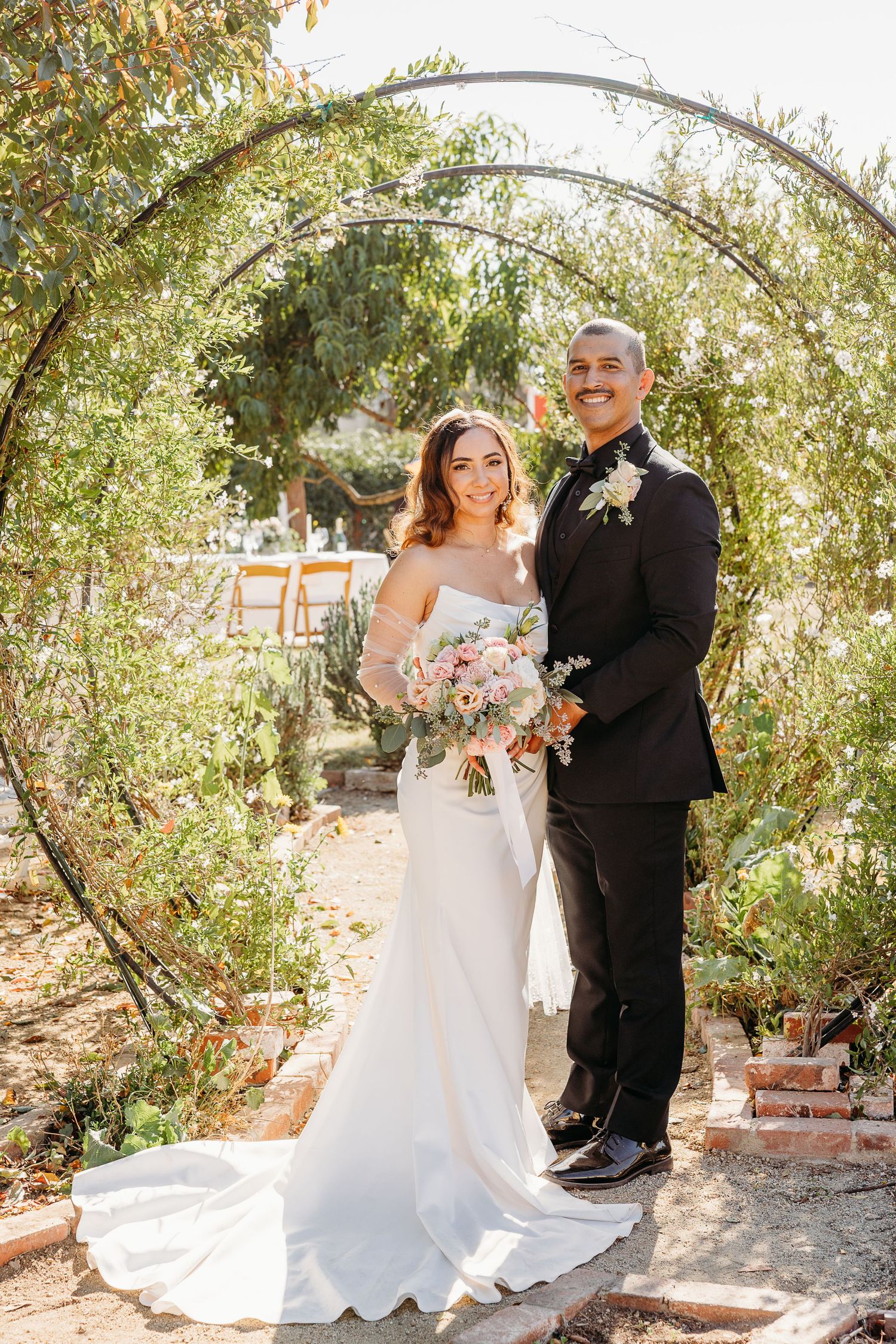 Wedding couple poses under natural archway of tree branches in outdoor garden setting.