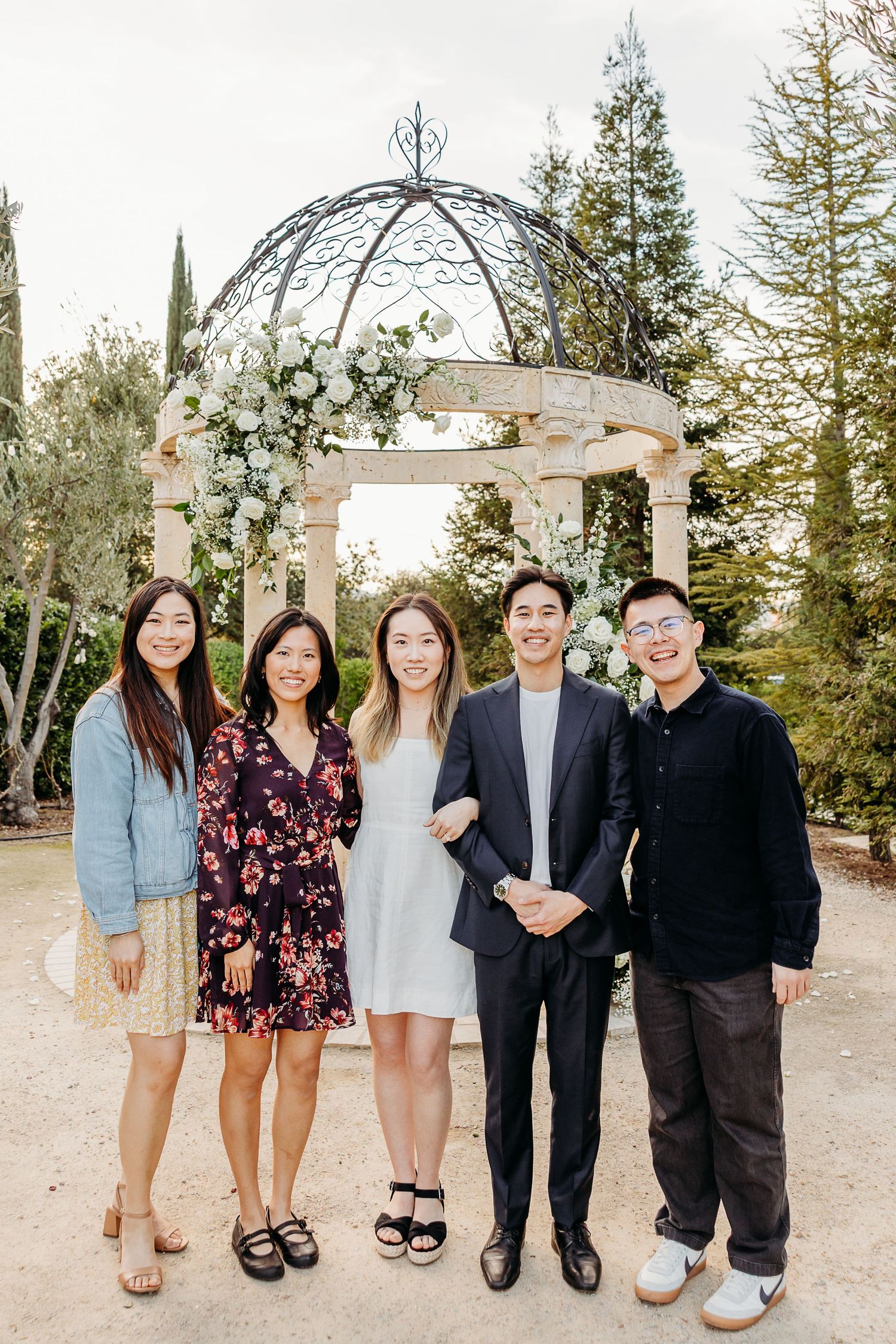 Wedding party posing together for the engagement photographer under a decorated white gazebo in an outdoor garden setting.