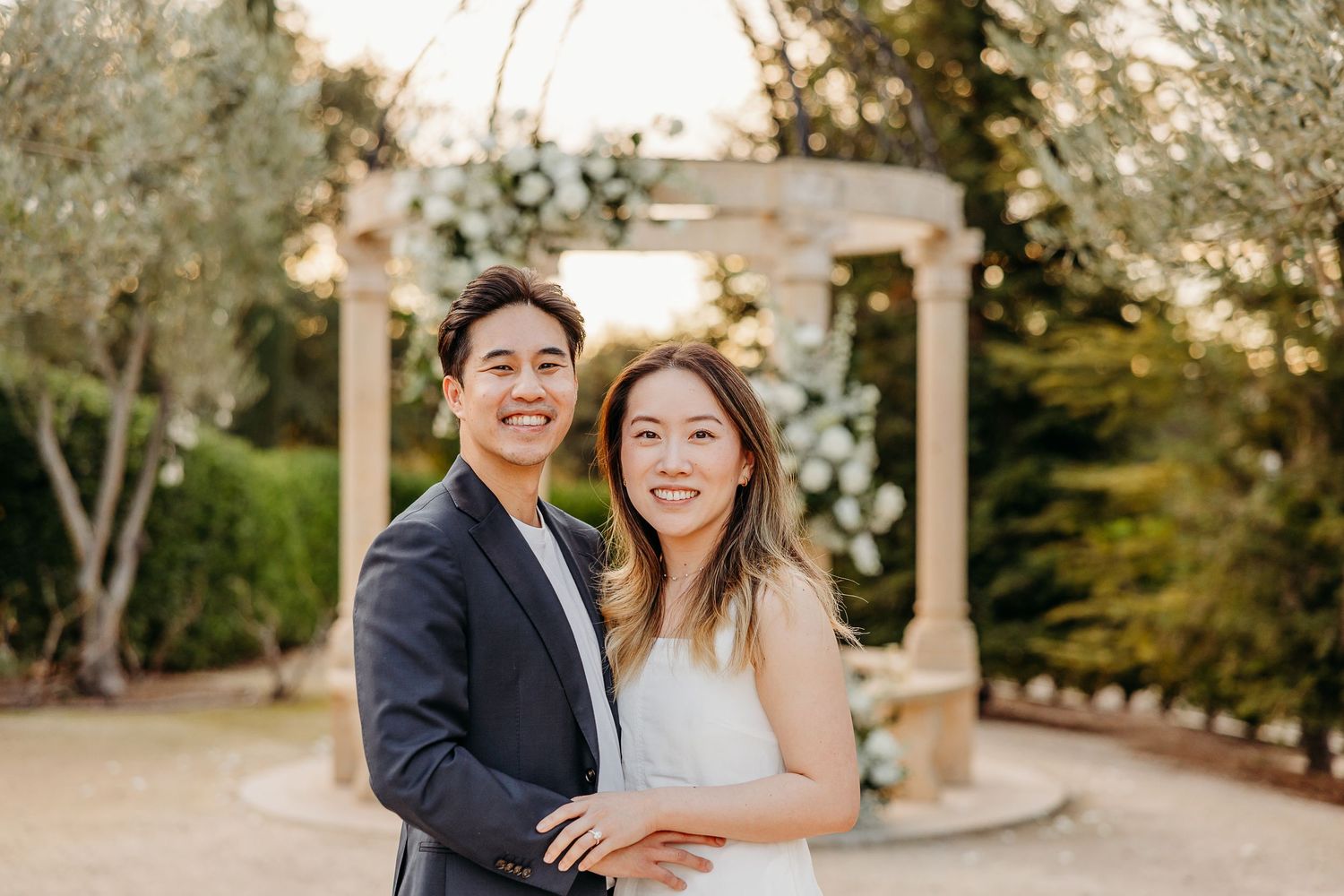 A joyful couple in wedding attire embrace and laugh together in front of a garden gazebo during golden hour.