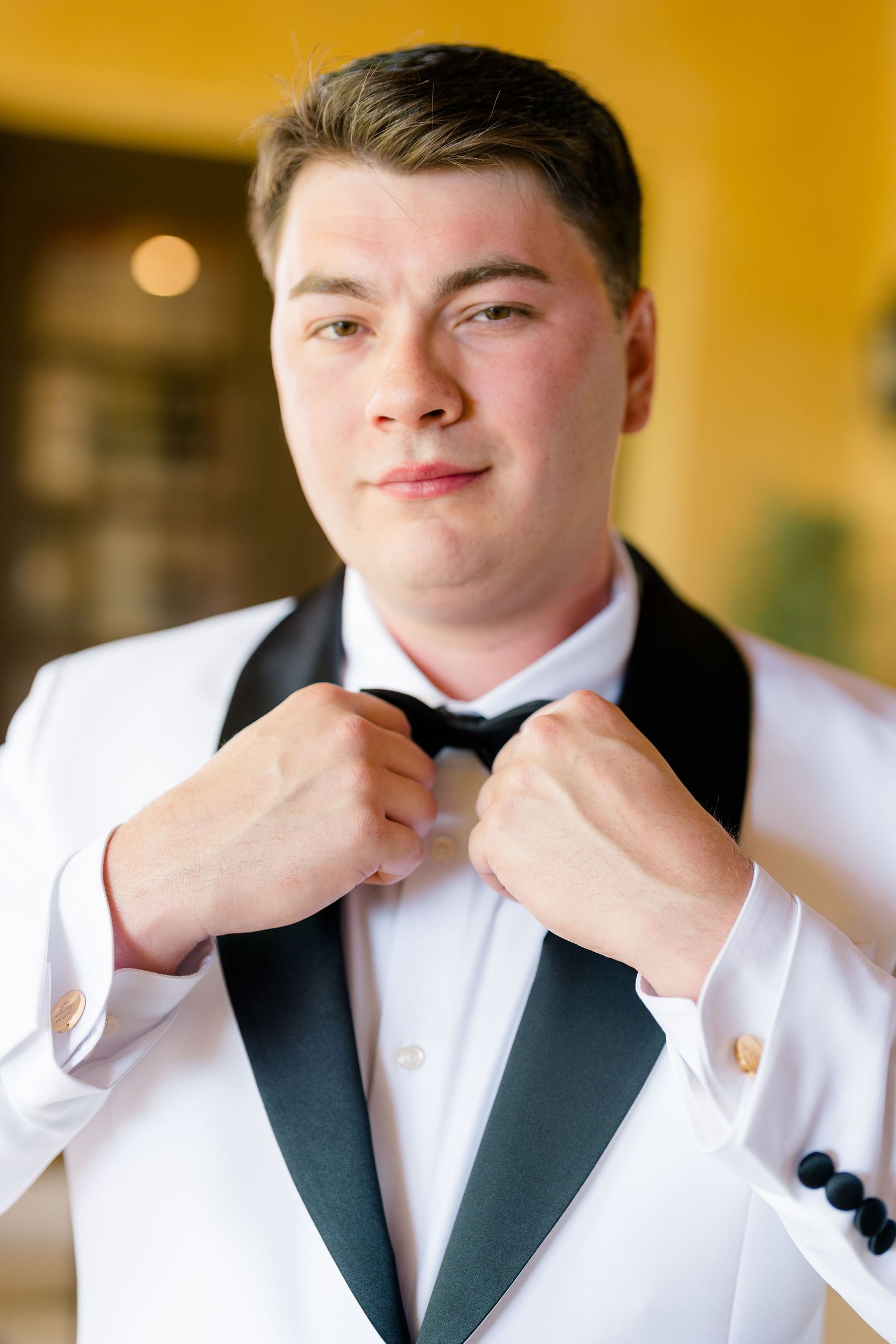 Young groom in white tuxedo jacket adjusting black bow tie, smiling confidently before wedding ceremony.