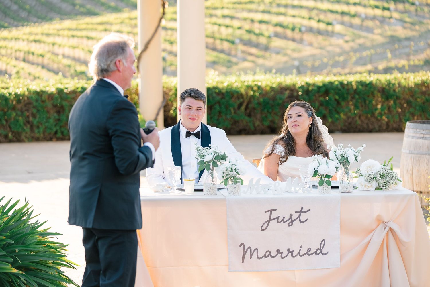 Newlyweds sit at Just Married sweetheart table as guest delivers speech at vineyard reception.