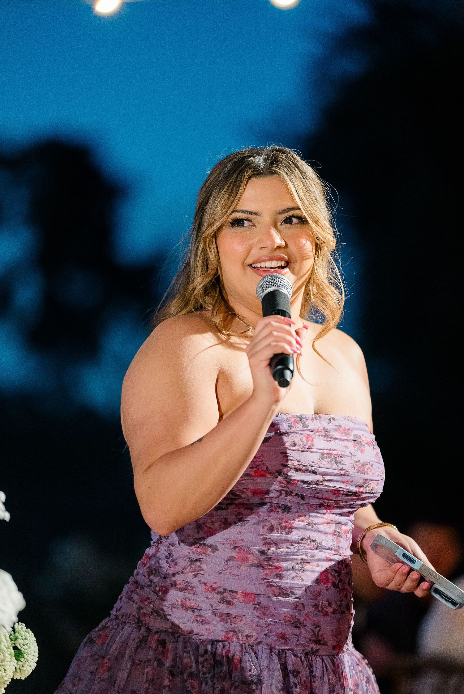 Blonde woman in floral strapless pink dress gives a speech at an outdoor evening wedding reception.