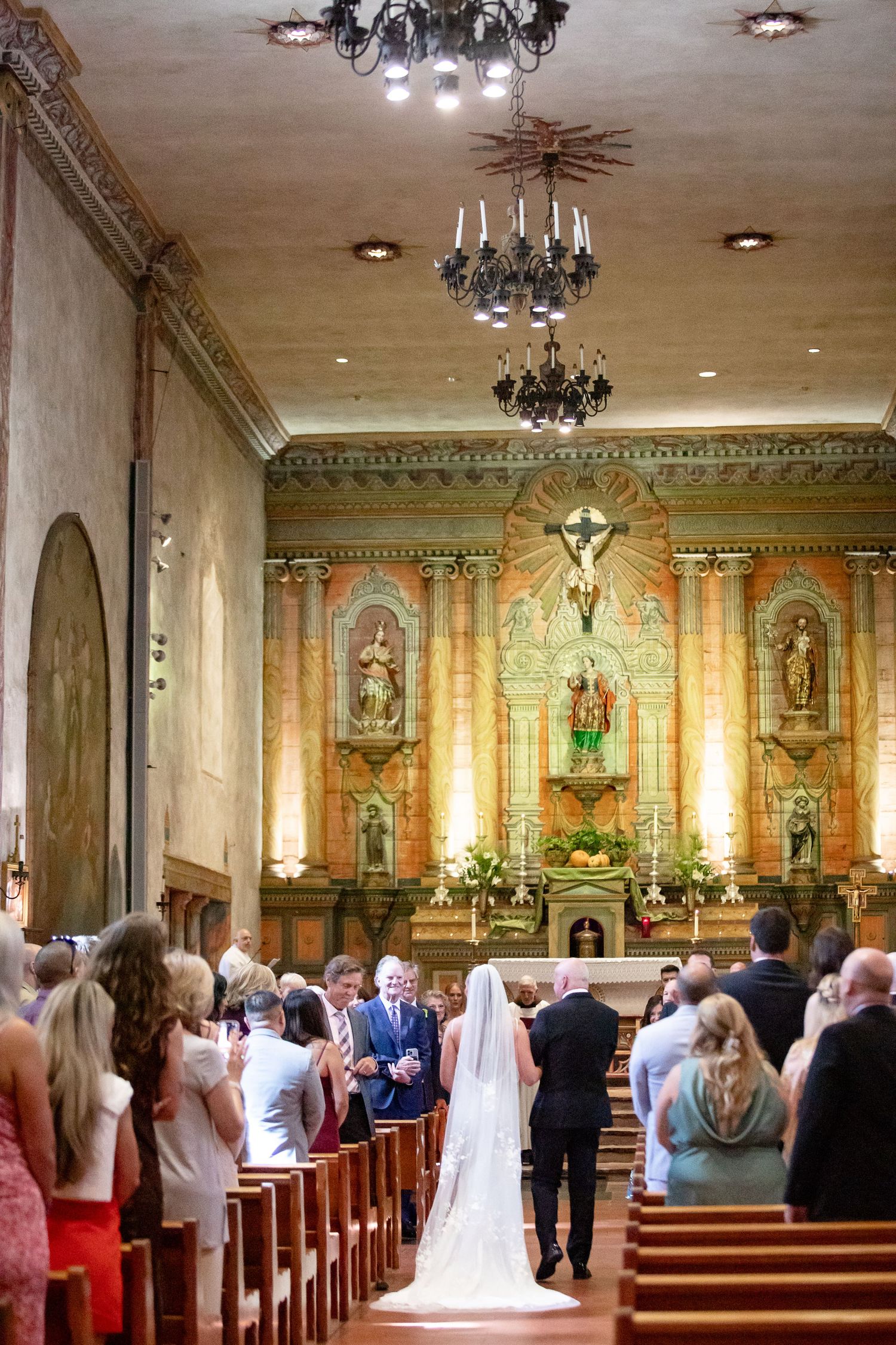 Wedding ceremony taking place in an ornate Catholic church with elegant chandeliers and religious artwork on display.
