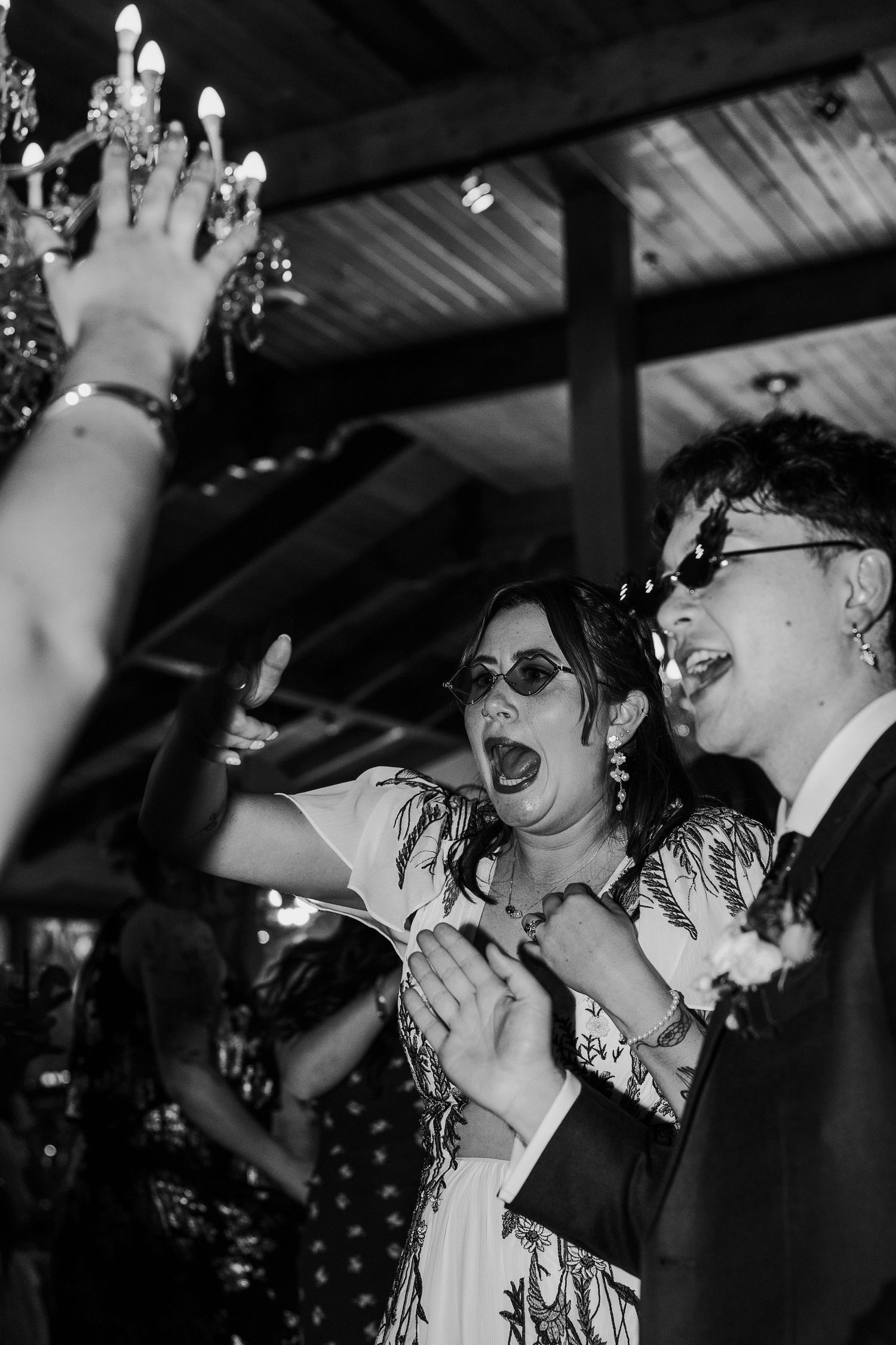 Black and white photo of guests dancing and celebrating at an indoor reception with chandelier lighting.