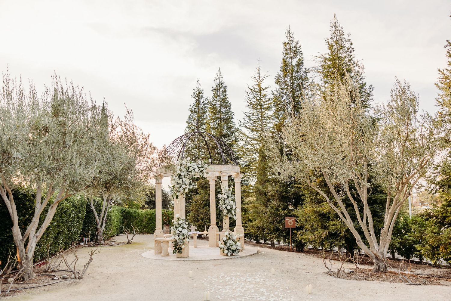 A white wedding gazebo with floral arrangements stands in a garden path surrounded by evergreen trees and bare branches.
