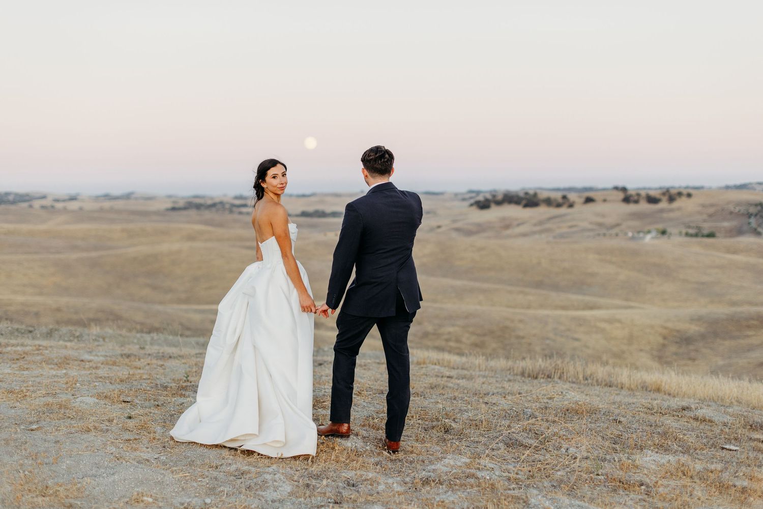 Couple walking hand in hand across a scenic hilltop landscape at golden hour.