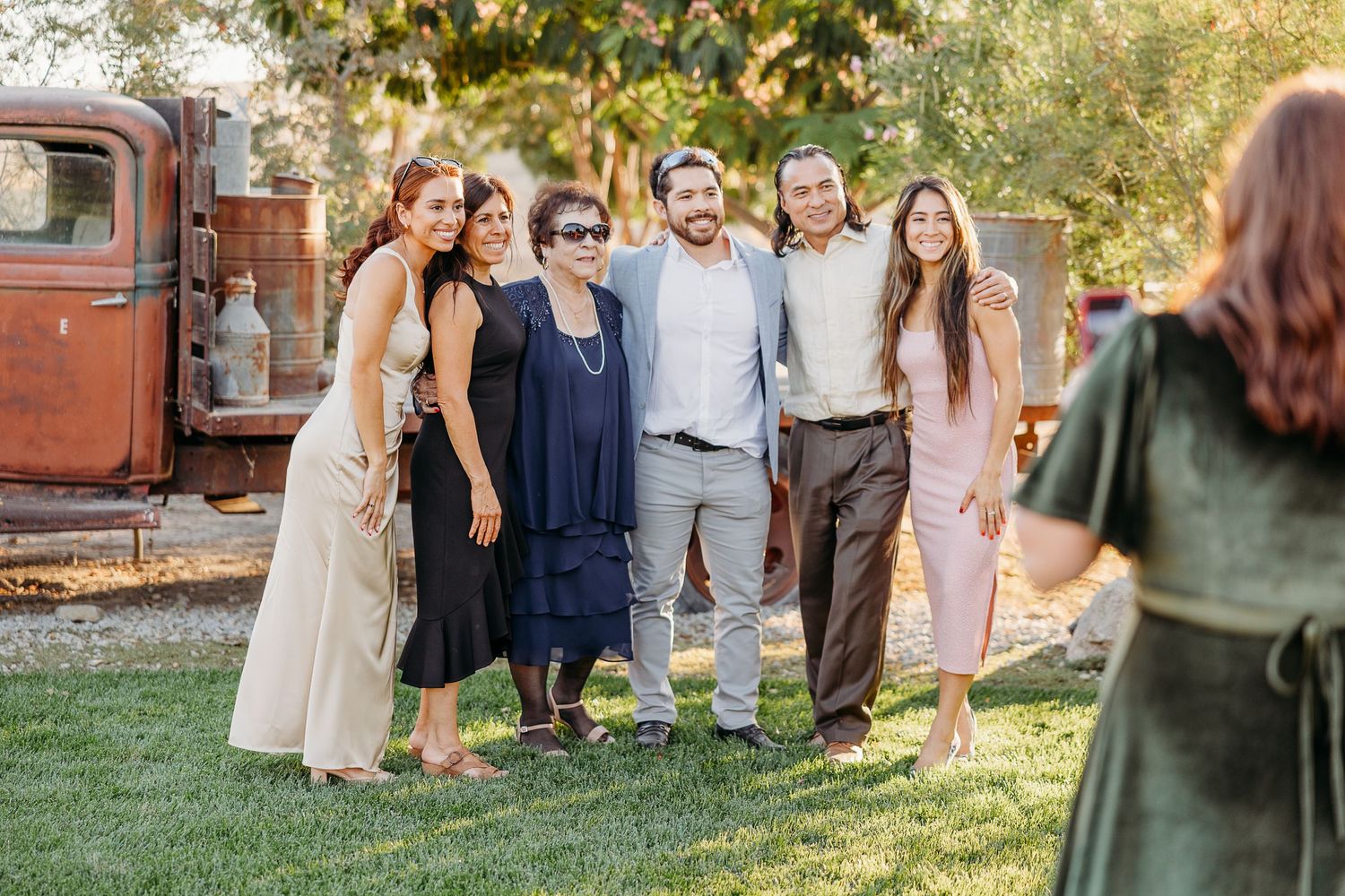 Wedding guests gather on a grassy lawn outside a rustic wooden barn venue during sunset.