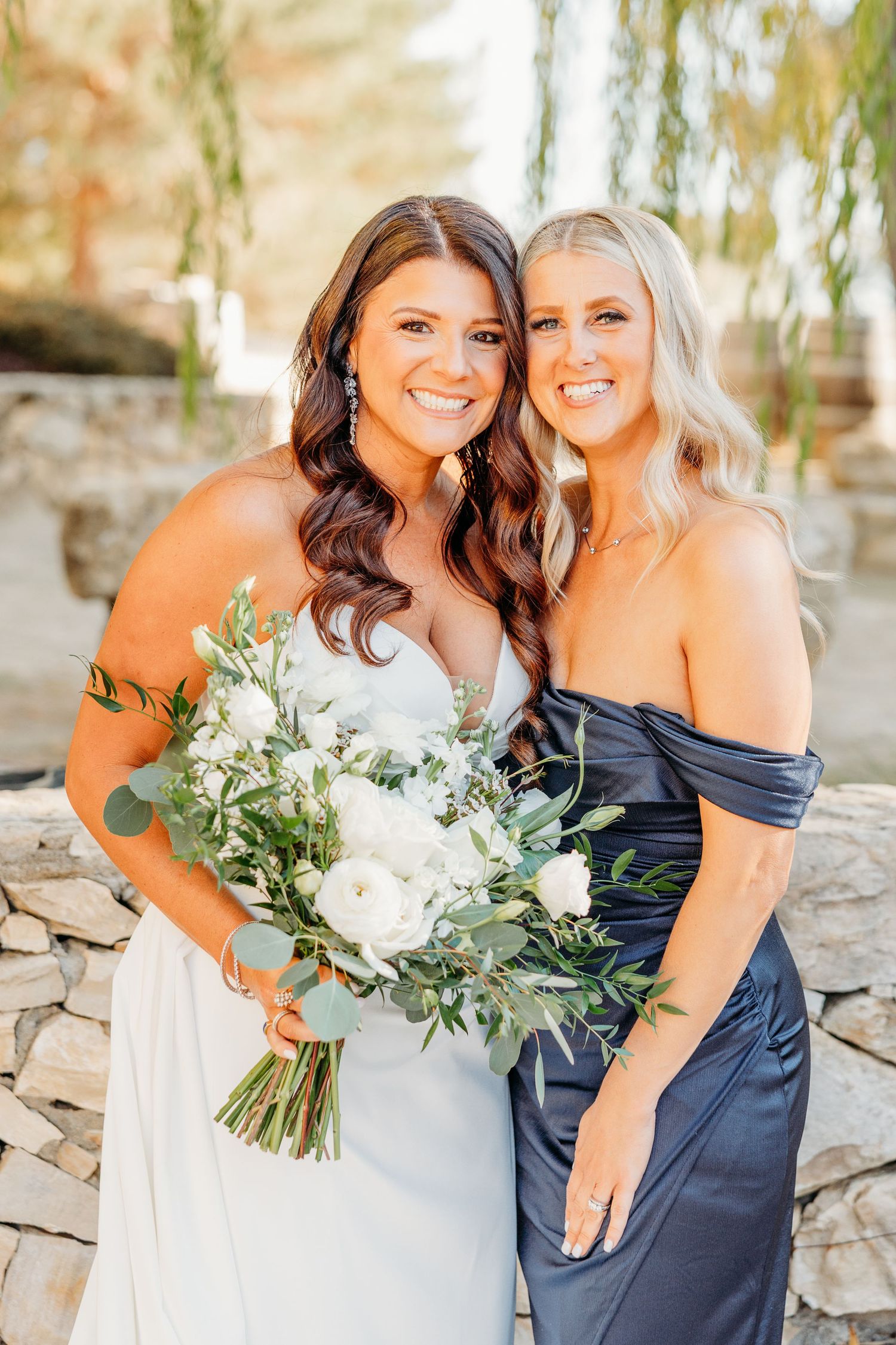 Wedding party members in navy dresses pose with elegant white floral bouquets against a stone wall backdrop.