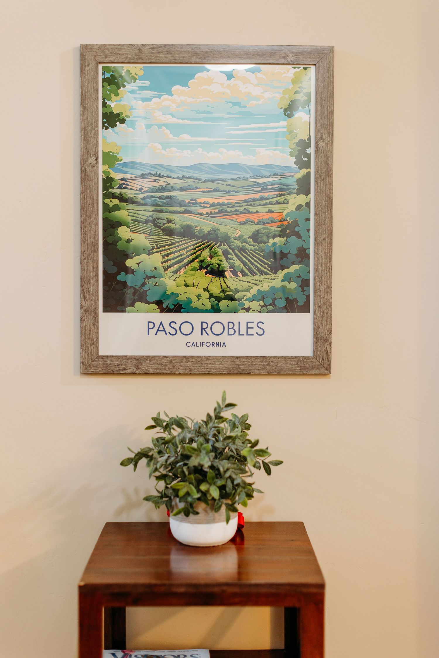 A framed landscape artwork of Paso Robles wine country with rolling hills and vineyards displayed above a wooden table with a plant.