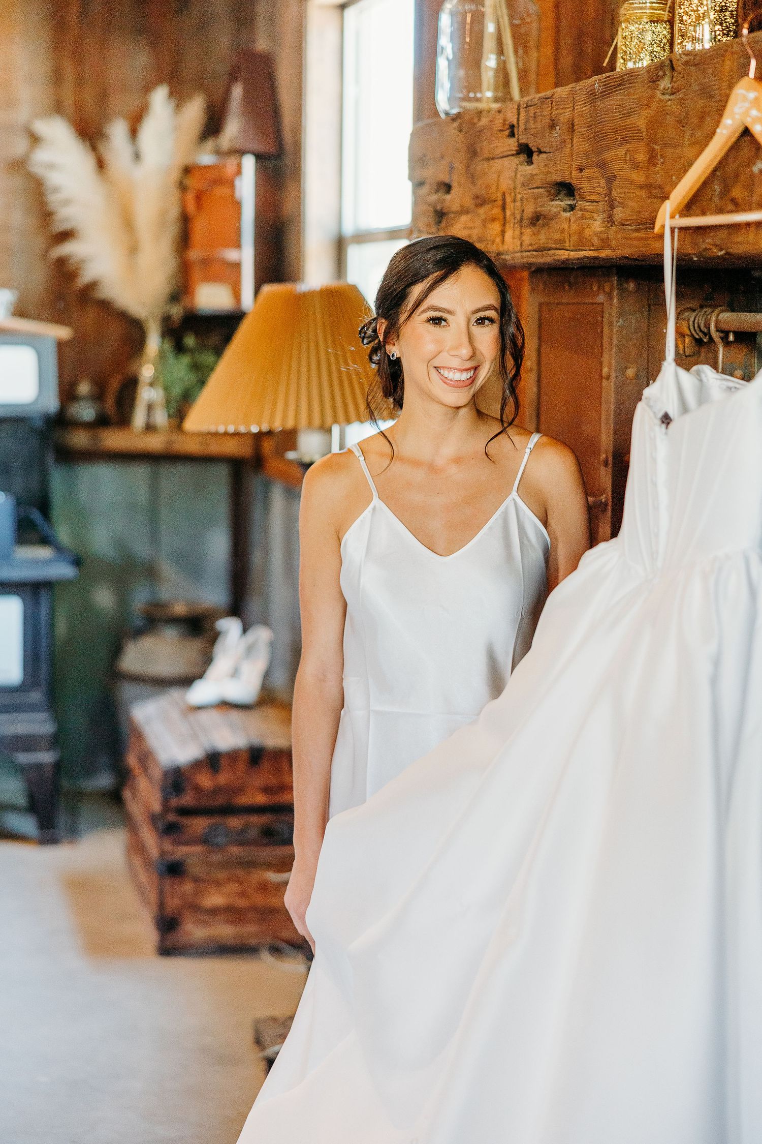 A wedding dress hangs in a rustic wooden barn with natural light streaming through windows.