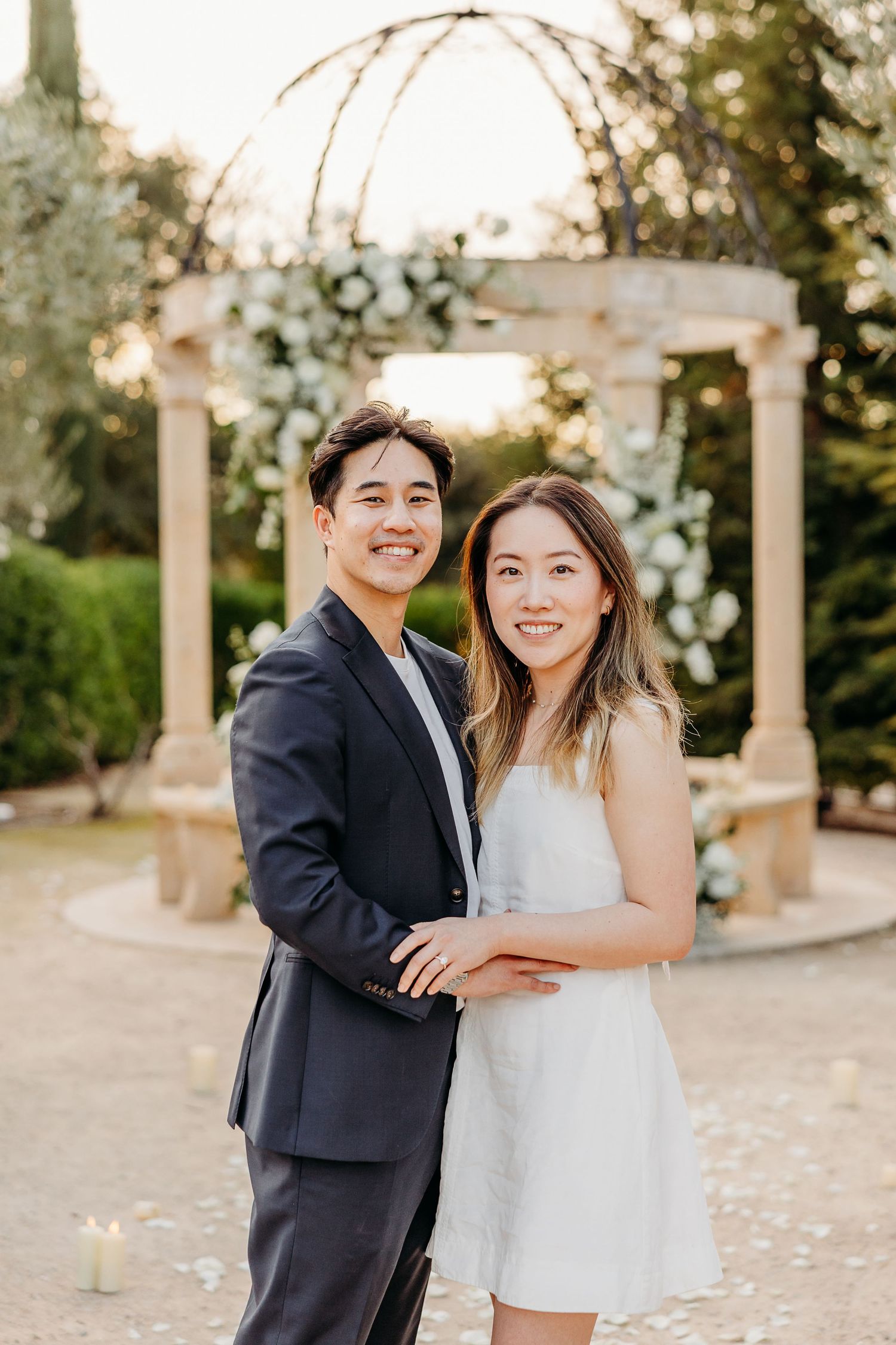 A couple poses together under a decorated garden gazebo with white floral arrangements.