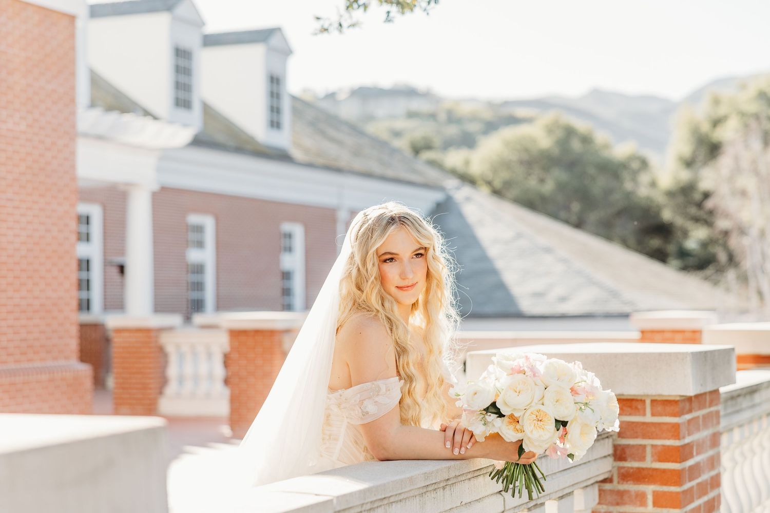 A bride in an elegant white gown poses on a brick balcony with mountain scenery in the background.