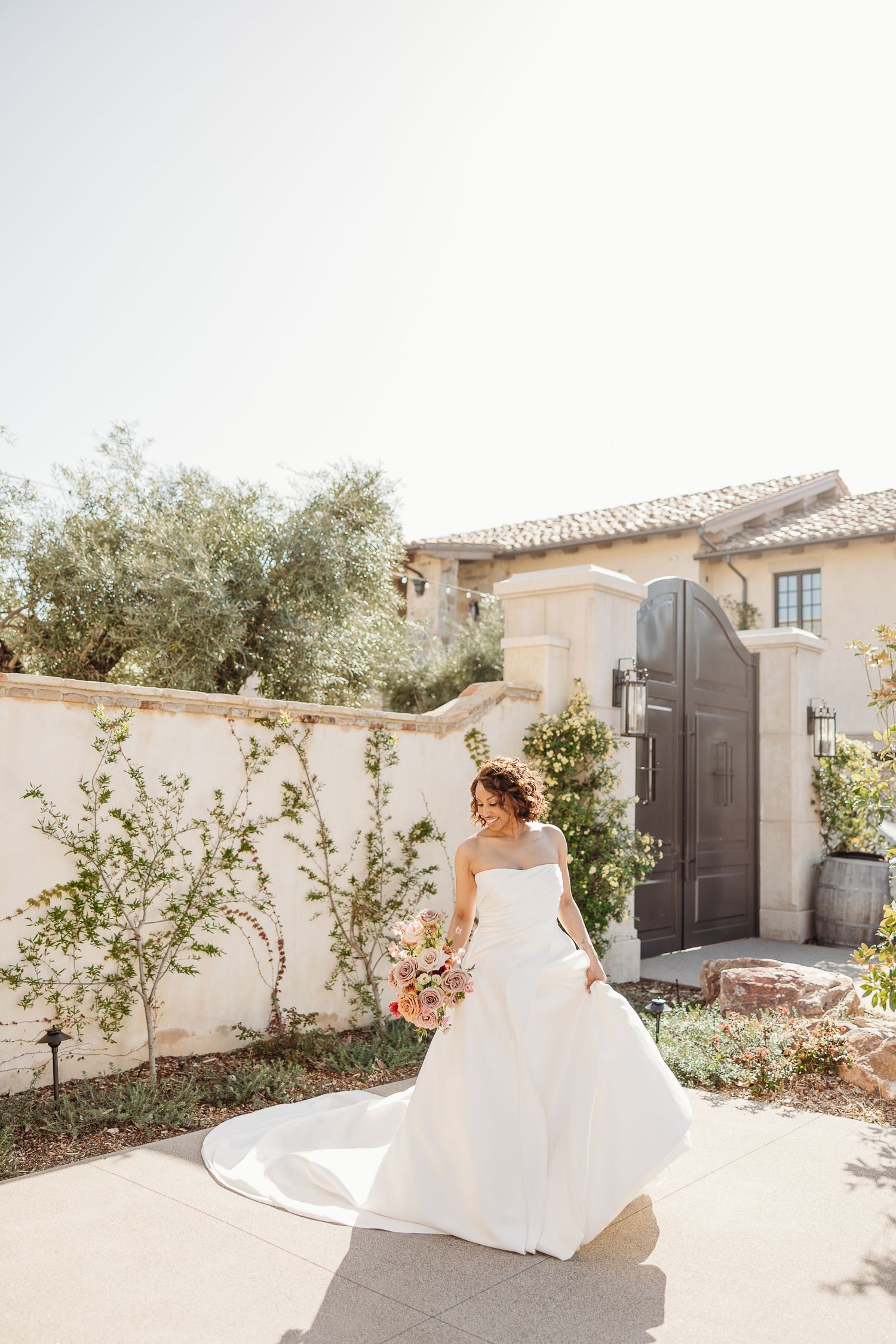 A bride in a flowing white wedding gown twirls gracefully in a sunlit Mediterranean-style courtyard.