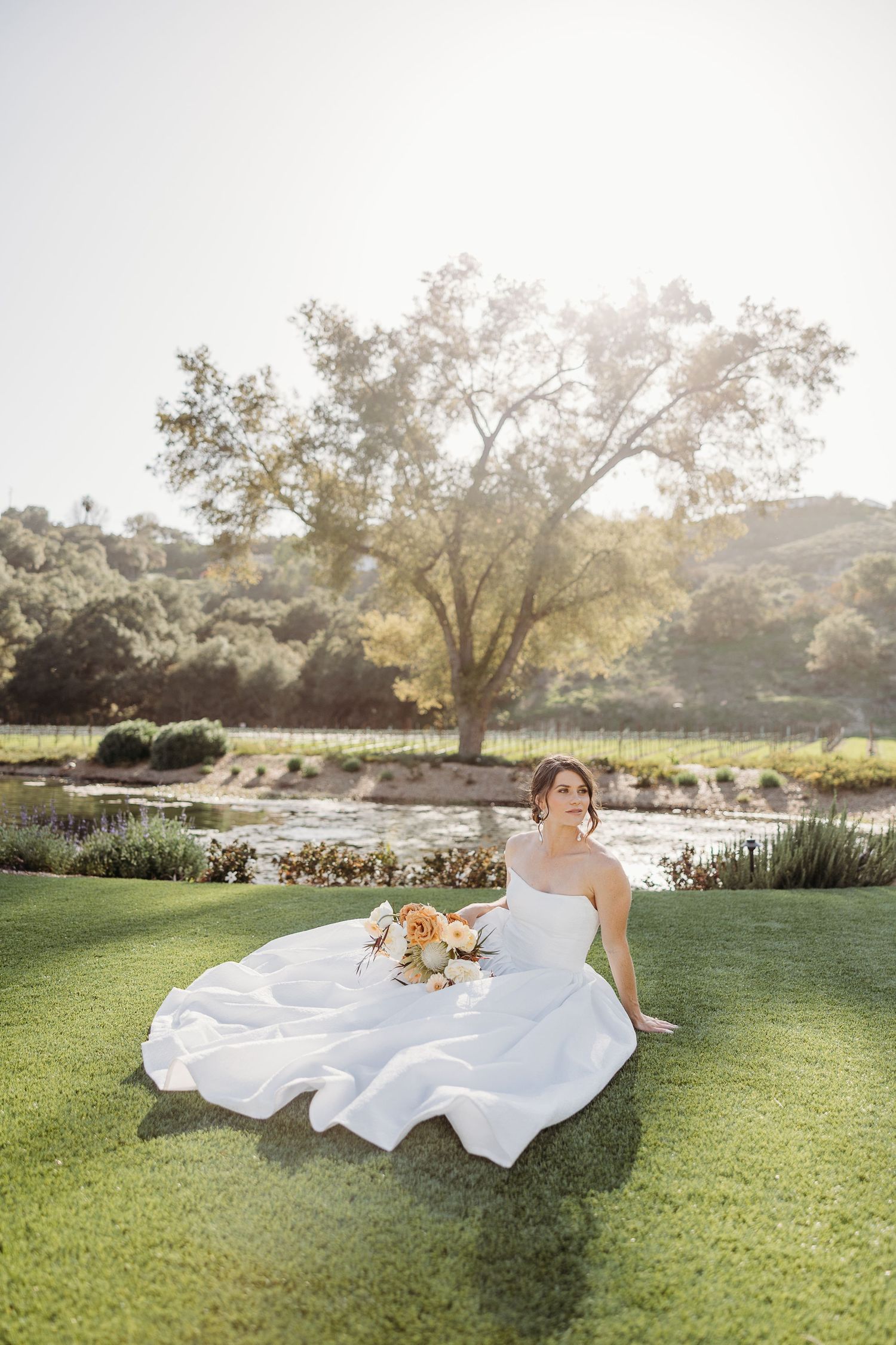 A bride in a flowing white dress lays gracefully on lush green grass during a sunset photoshoot at an outdoor venue.