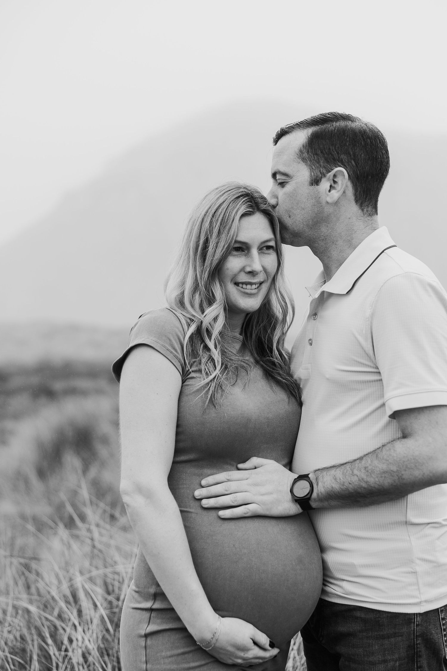 Black and white maternity photo capturing couple's joy in coastal setting.
