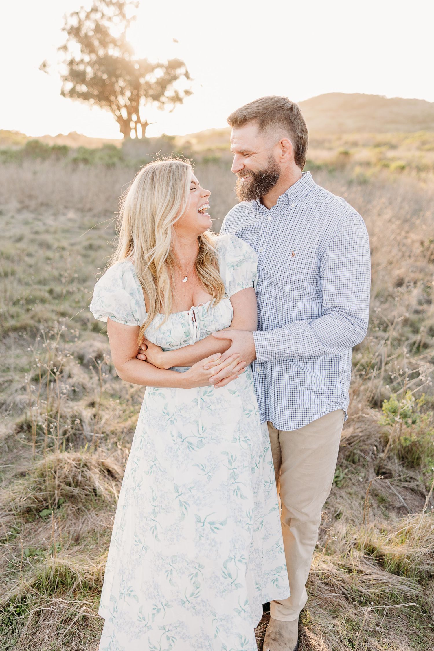 A couple shares a joyful moment together in a grassy field at sunset during a romantic outdoor photoshoot.