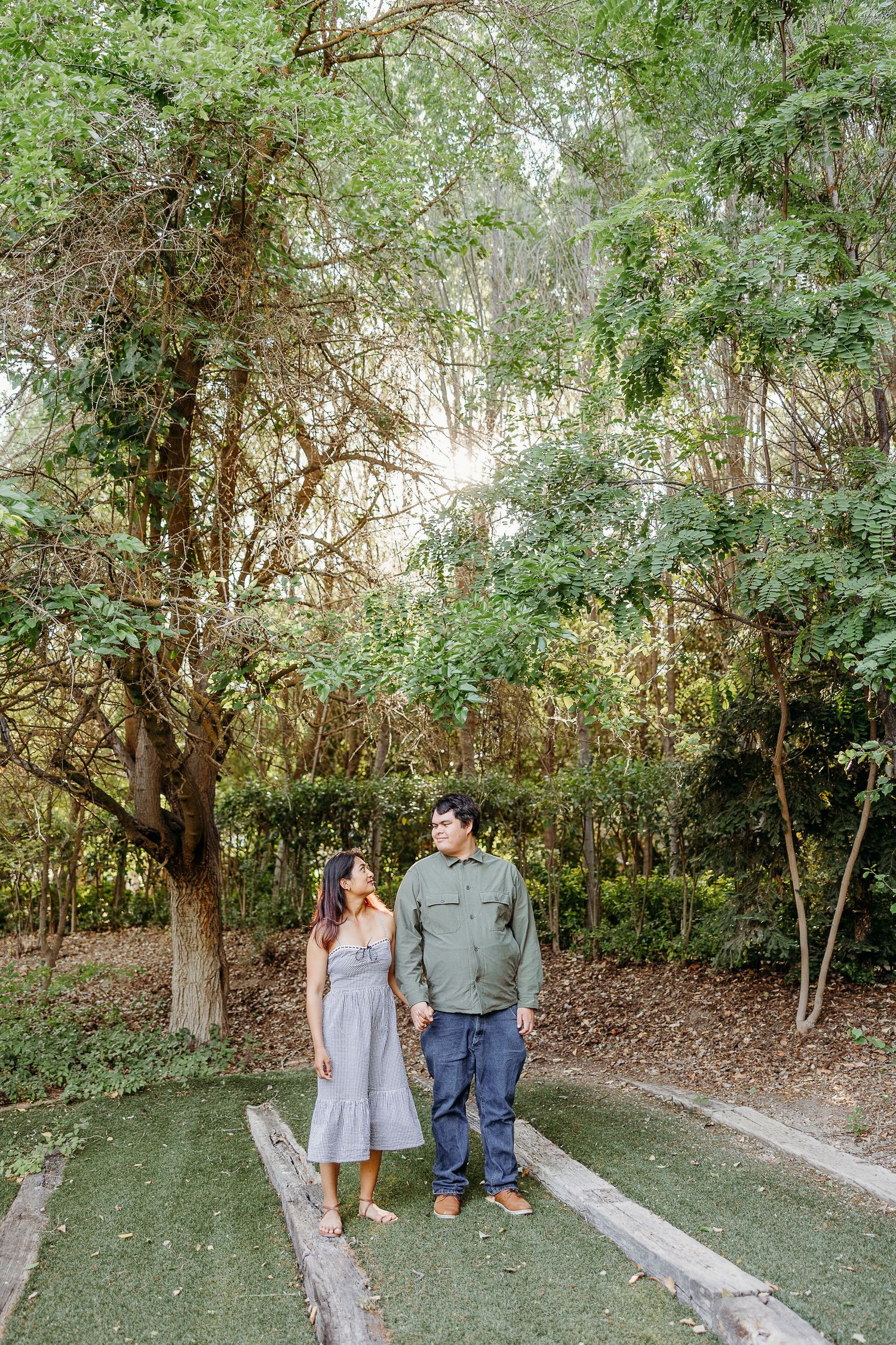 Two people share a loving moment while walking along a tree-lined path in a natural outdoor setting.