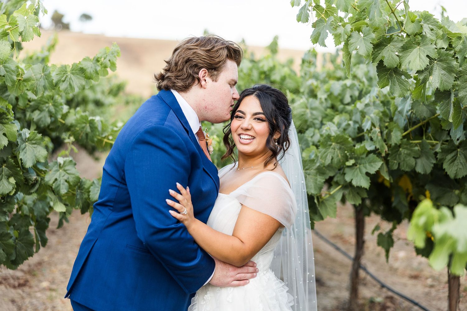 Happy couple embrace and laugh together among vineyard rows on their wedding day.