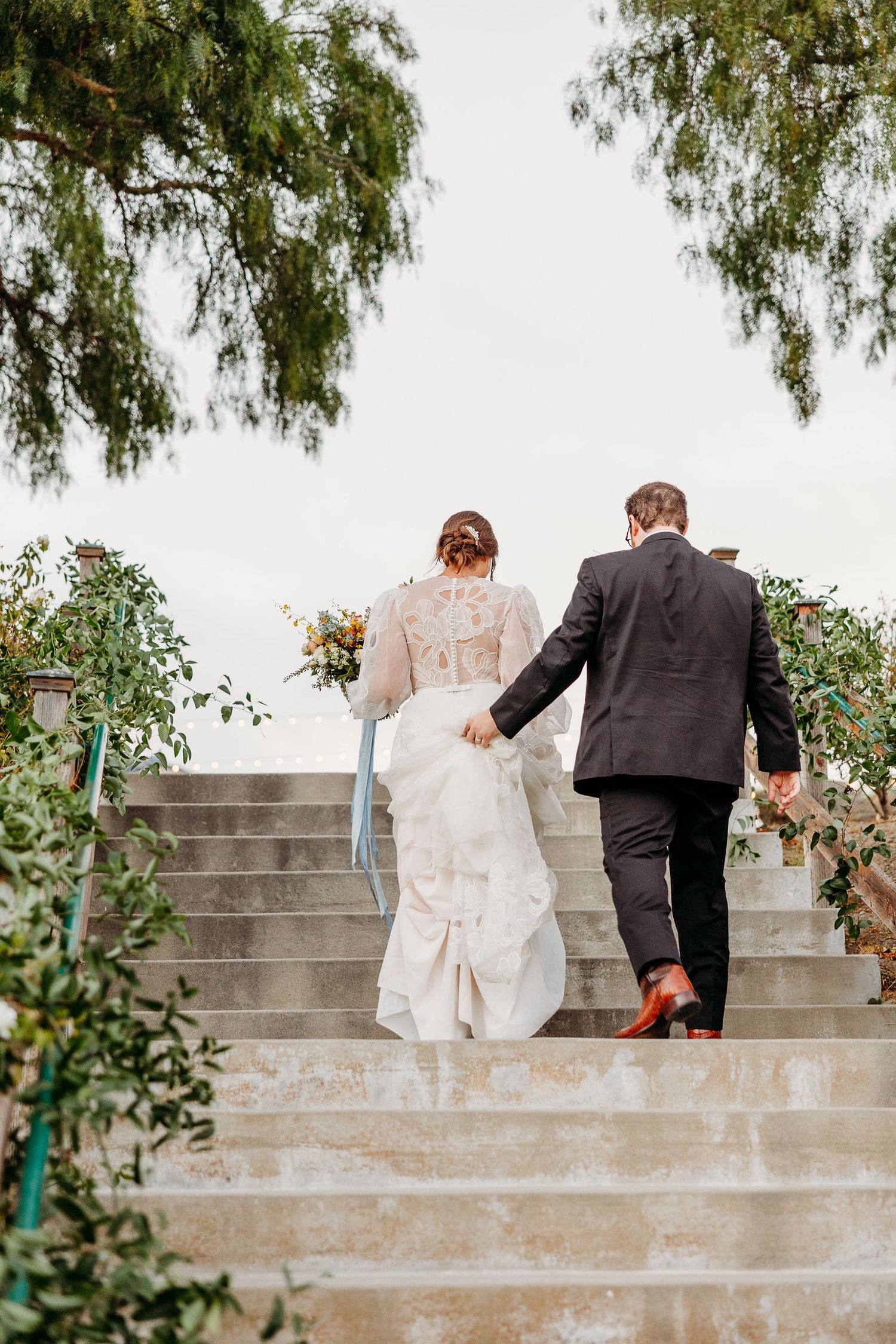 Newlywed couple ascending stone steps together, with the bride holding a bouquet and wearing a flowing dress with blue accents.
